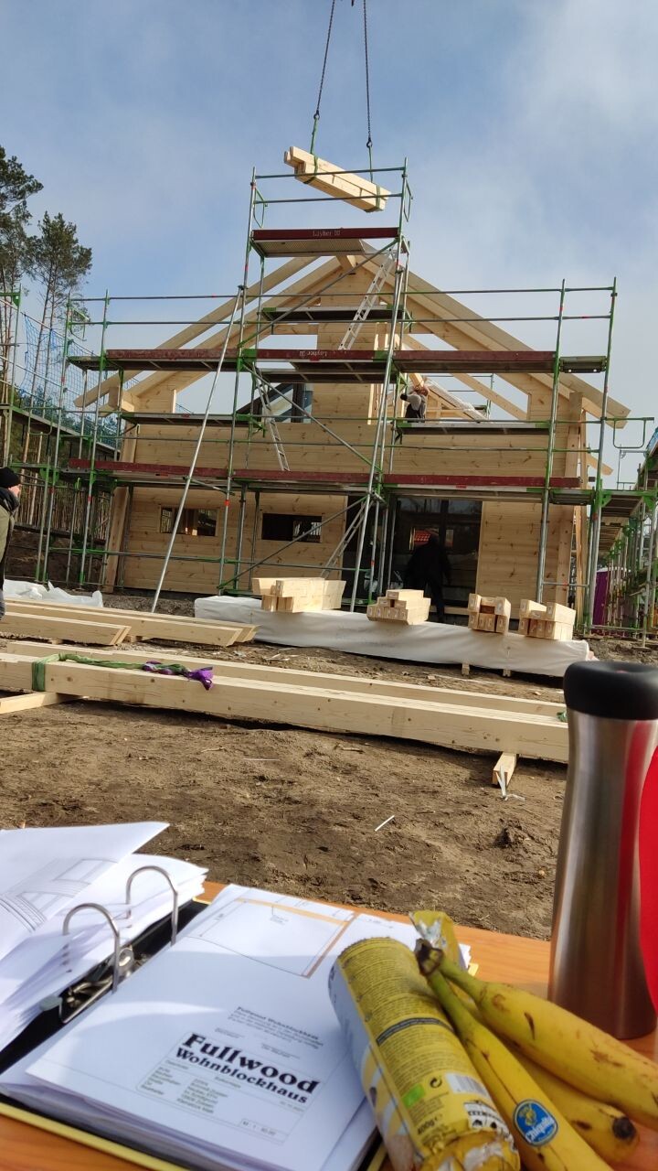 Holzhausbau mit Gerüst, Kran hebt Holzbalken; Baupläne liegen auf dem Tisch