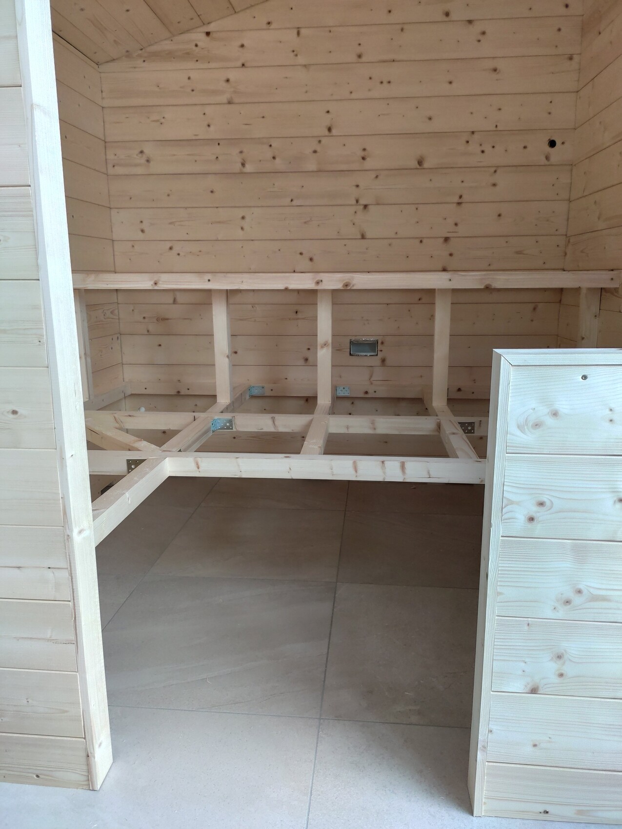 Innenraum eines Holzhauses mit freiliegendem Holzrahmen an der Wand und Fliesenboden.