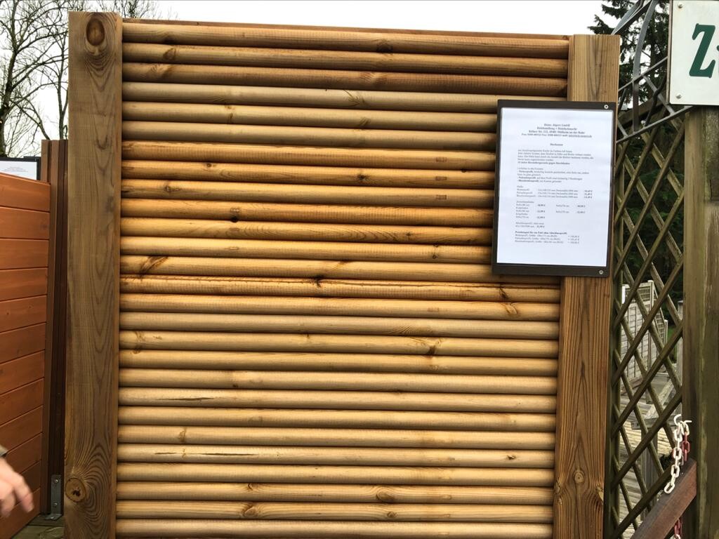 Holzlattenwand mit rechts montierter Infotafel; Zaunstruktur im Hintergrund