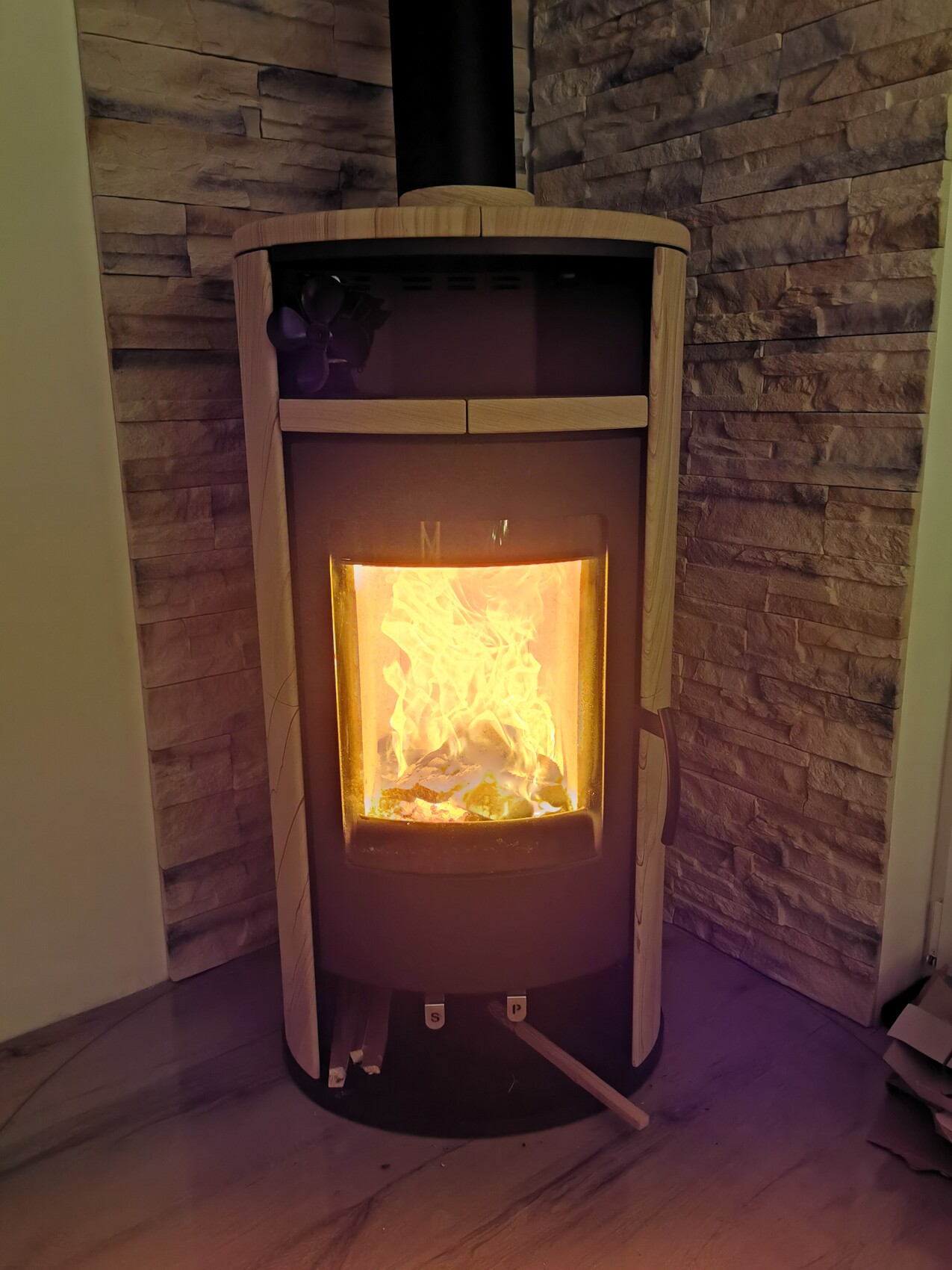 Holzofen mit Glastür brennt Flammen vor Steinwand-Verkleidung, gemütlicher Kaminraum.