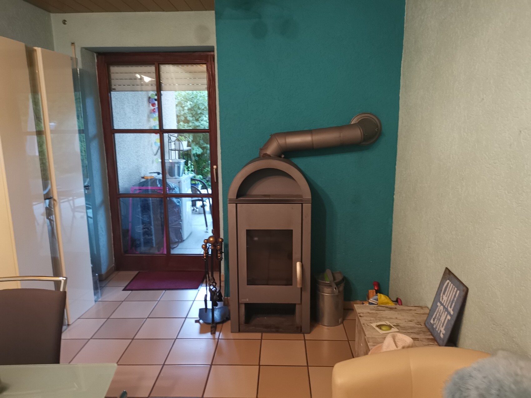 Ein grauer Kaminofen mit Rohrbogen an türkisfarbener Wand, Glastür nach außen.
