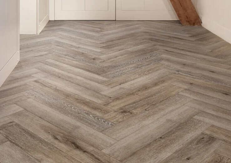 Heller Holzlaminatboden im Fischgrätmuster in einem Innenraum mit Türen im Hintergrund.