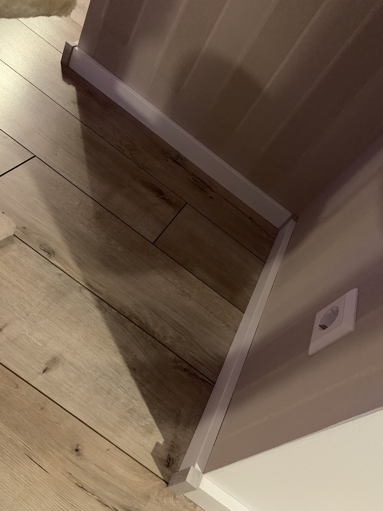 Ecke mit Holzfußboden, weißer Sockelleiste und Wandsteckdose.