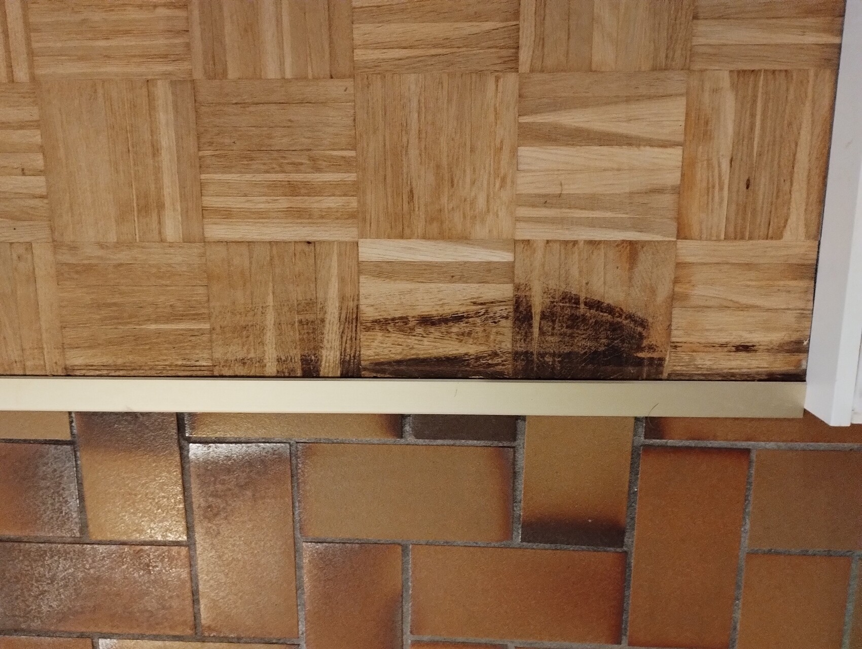 Holzparkettboden mit dunklem Fleck unten am Rand, Übergang zu braunen Terrakottafliesen.
