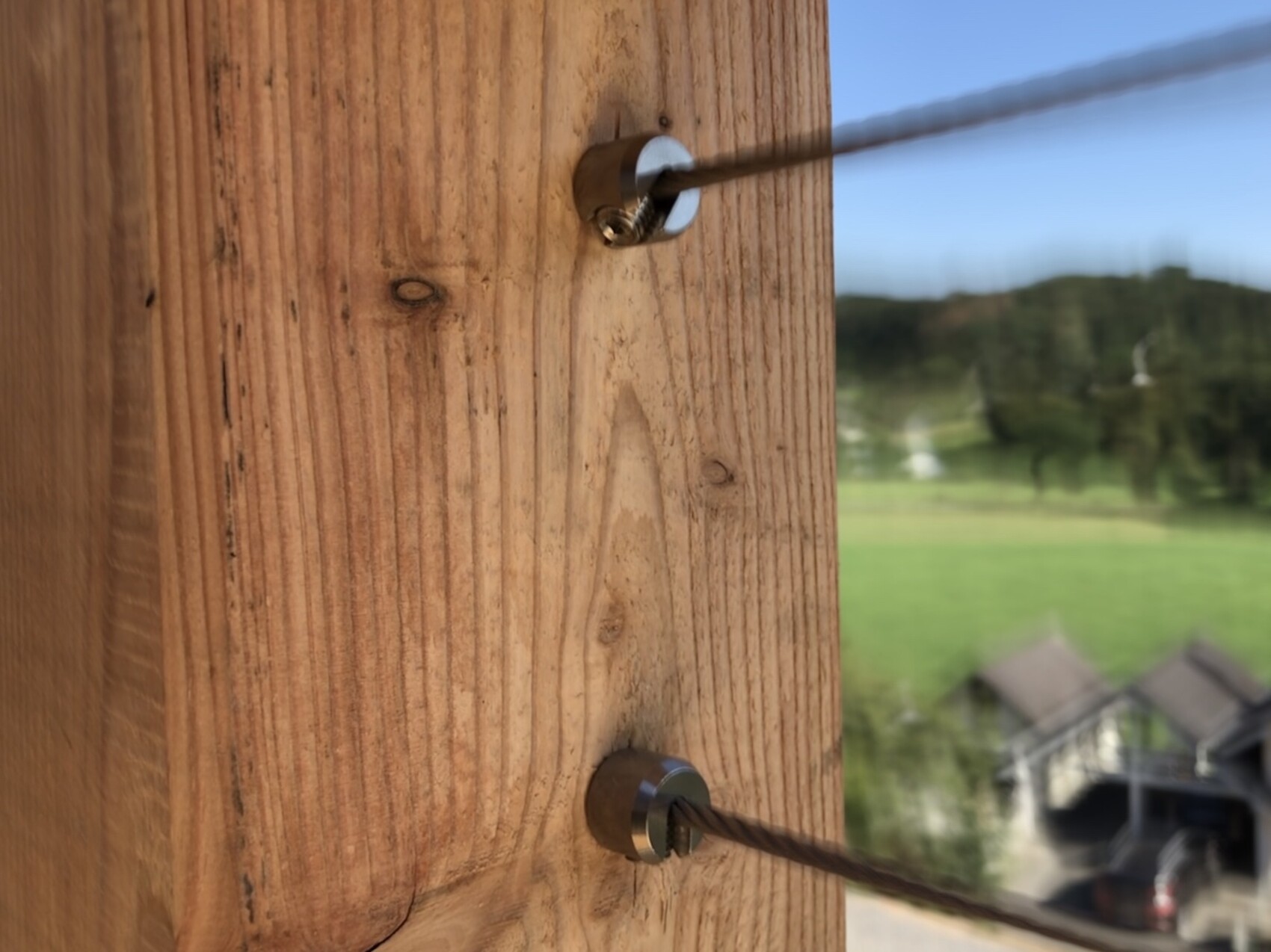 Nahaufnahme eines Holzpfostens mit Edelstahlseil und Spanner, Landschaft im Hintergrund