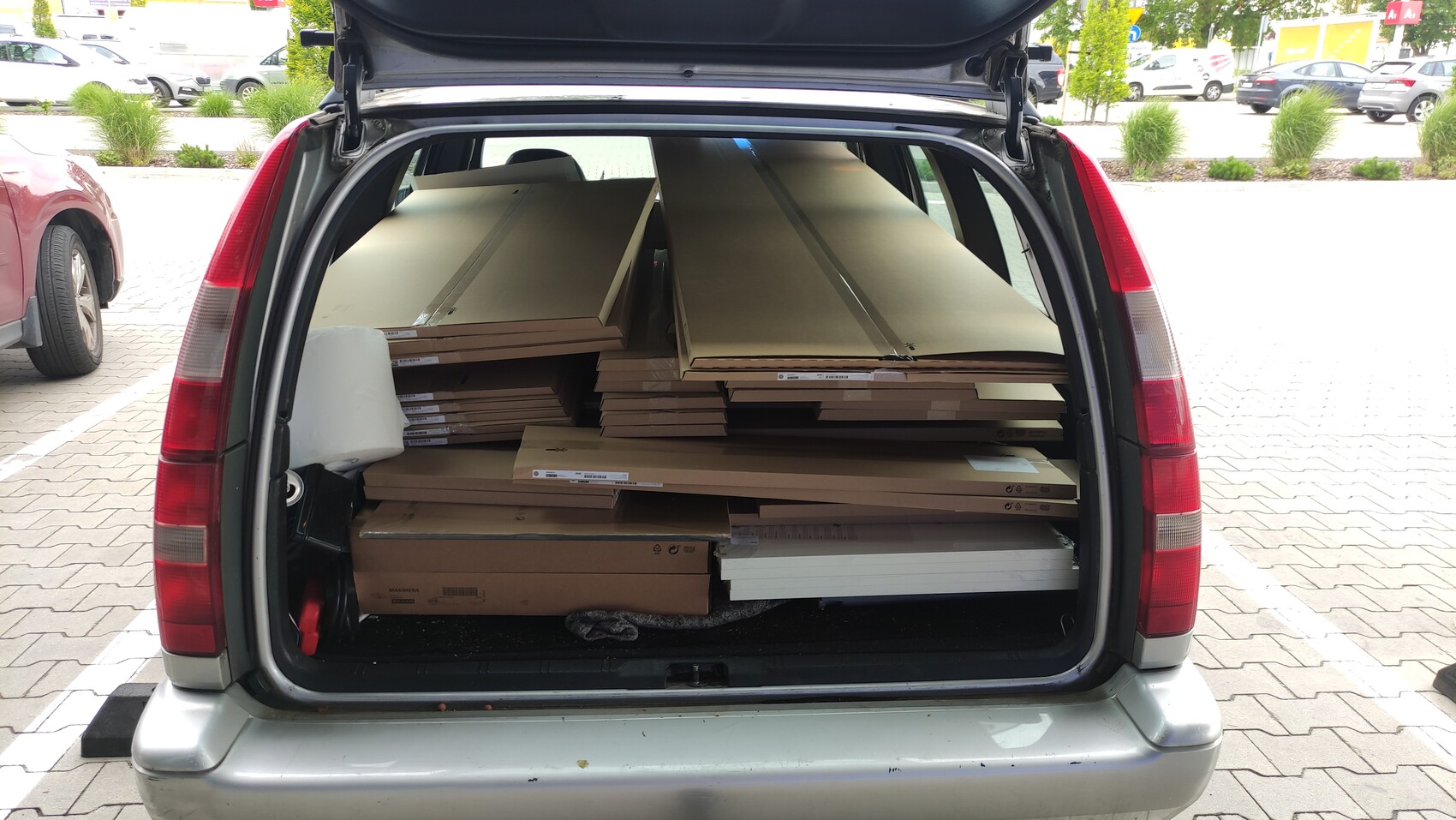 Offener Kofferraum eines silbernen Autos mit gestapelten Holzplatten und Kartonverpackungen.