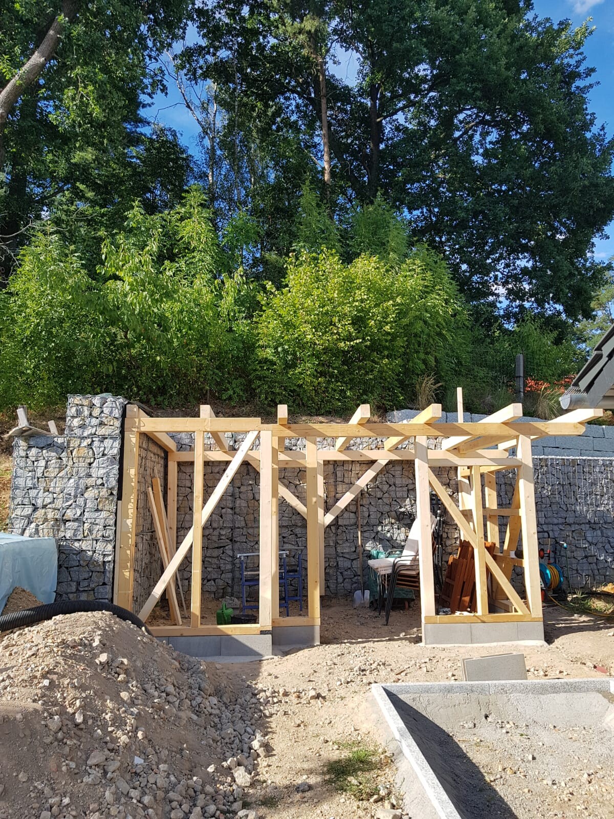 Holzrahmenbau vor einer Stein-Gabionenwand; Baugerätschaften und Erdreich sichtbar.