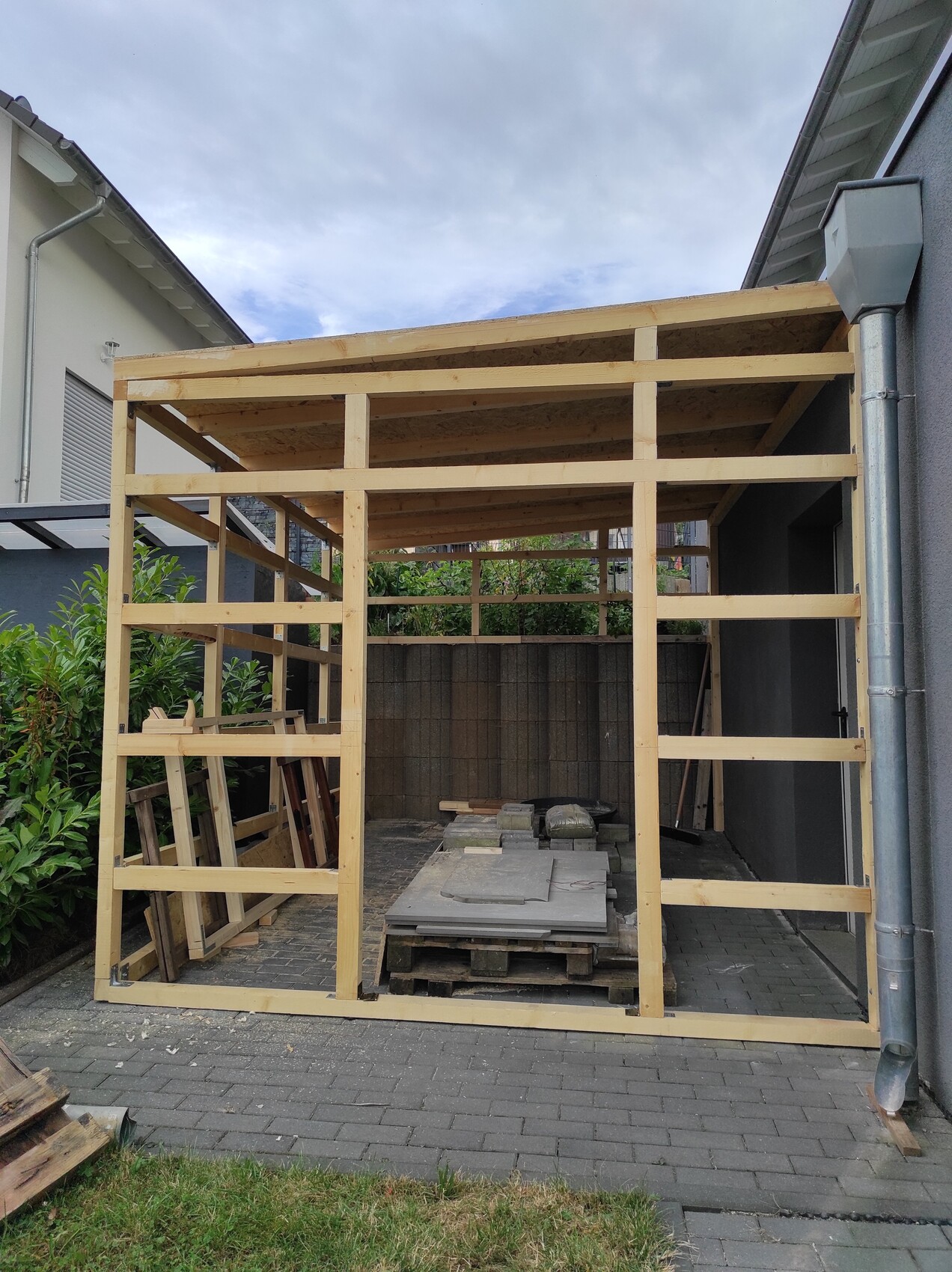 Offene Holzrahmenkonstruktion eines Garten-Sheds im Bau, mit Paletten und Baumaterialien.