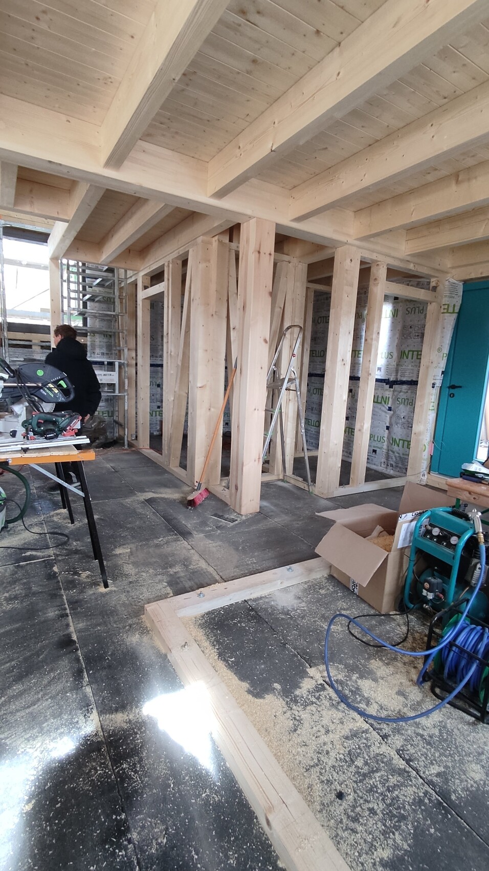 Innenraum einer Baustelle: Holzständerwerk, Dachbalken, Leiter und Werkzeuge, Person mit Säge.