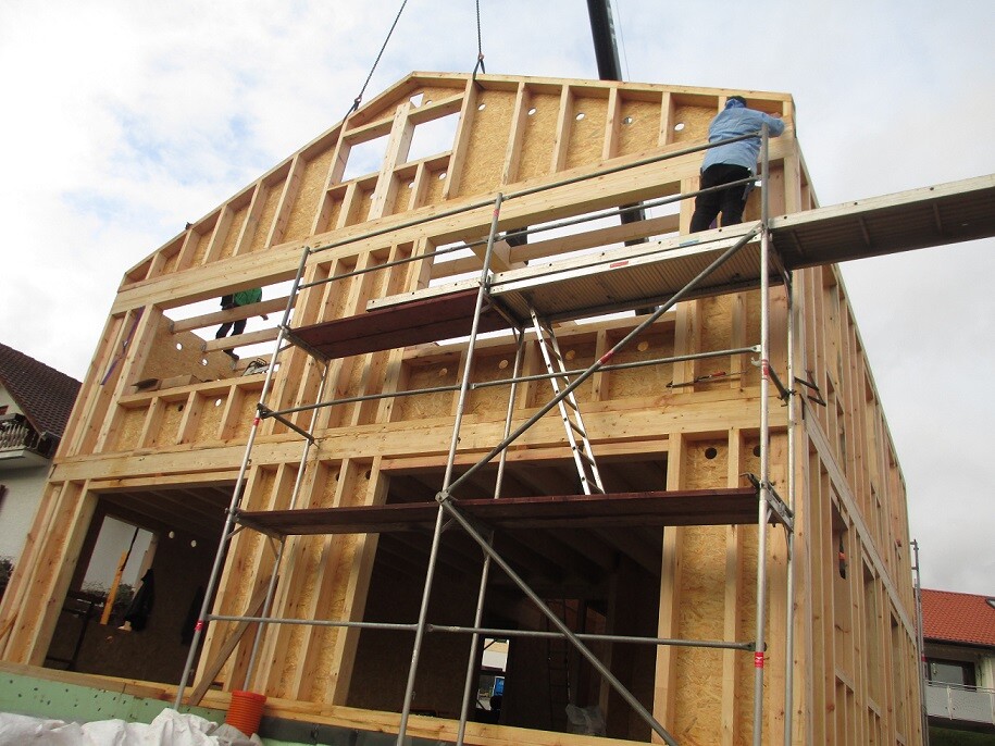 Rohbau eines Holzhauses mit Gerüst, Arbeiter oben am Dachbereich, Kranelement sichtbar