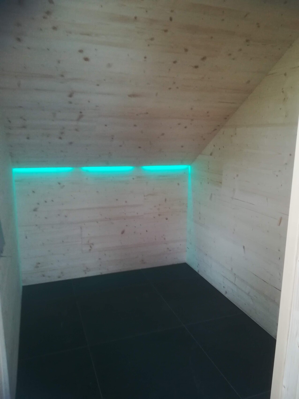 Kleiner Raum mit Holzwänden und Decke, dunklem Boden; türkis LED-Lichtleiste an der Wand.