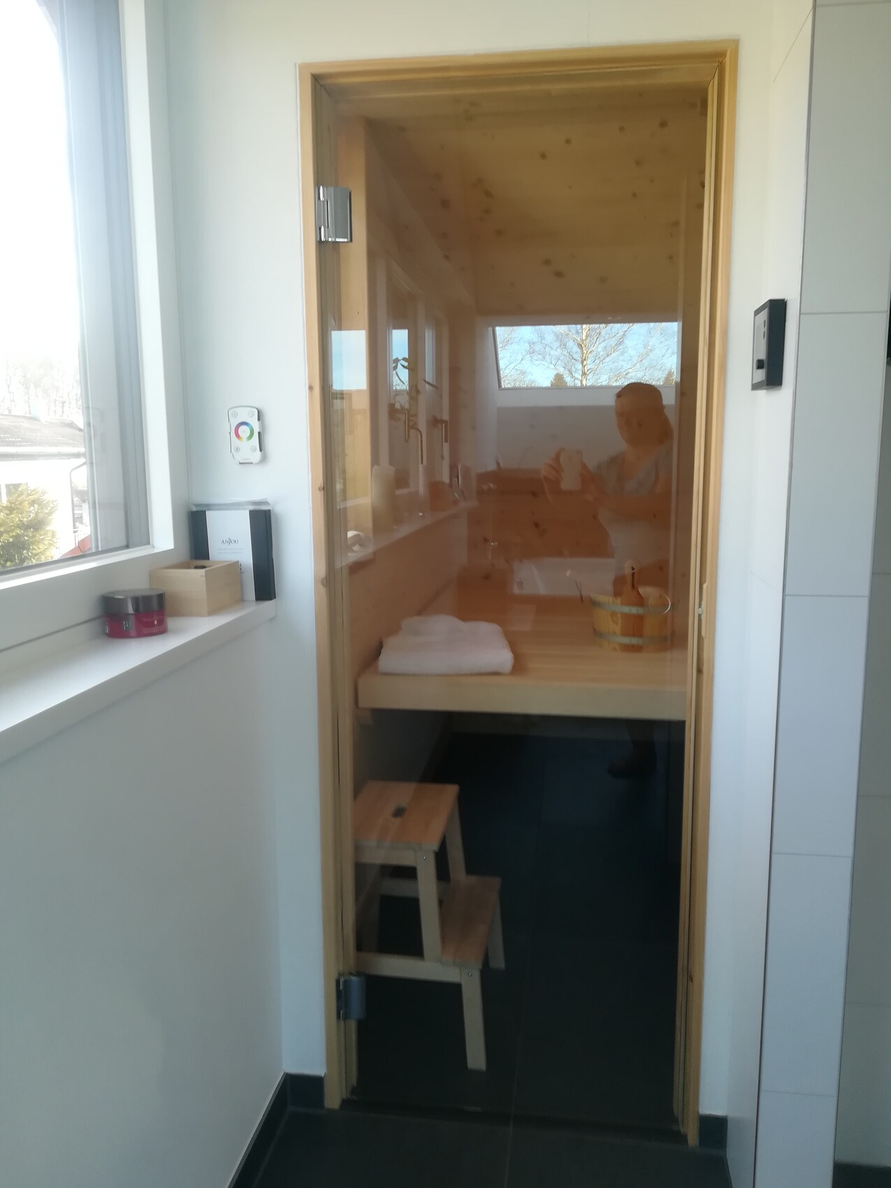 Holzsauna mit Bank, Handtuch und Eimer; Person fotografiert durch die Glasfront.