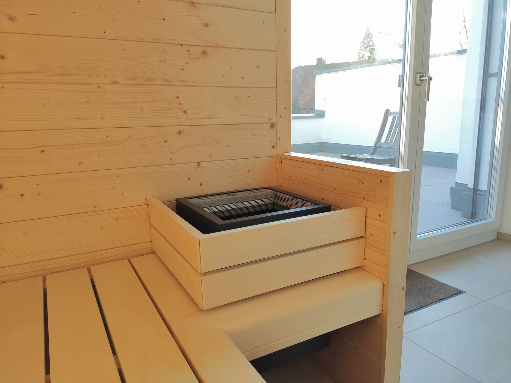 Heller Holzsauna-Innenraum mit Holzbank, Ofen in Holzkiste und Tür zur Terrasse.