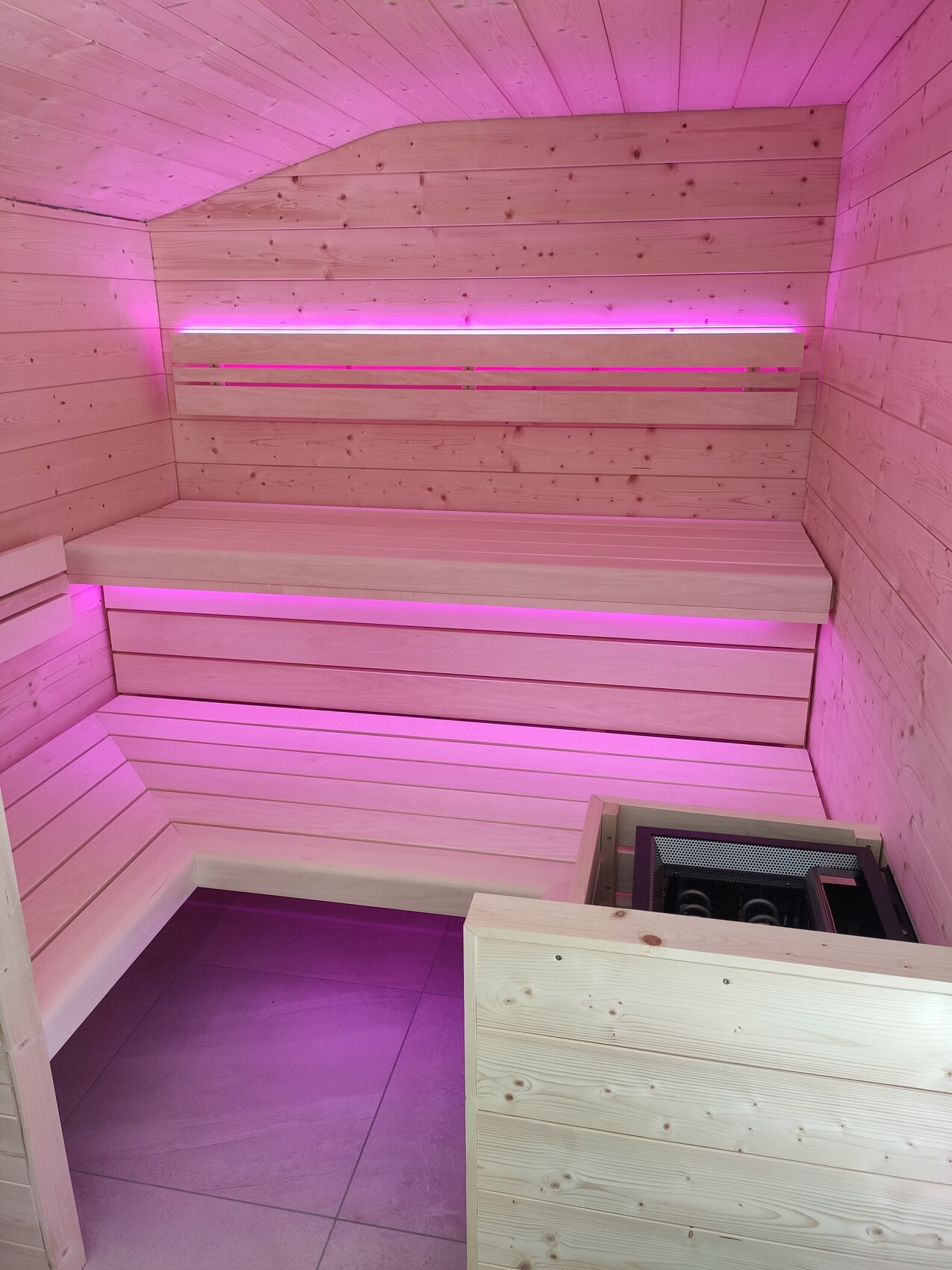 Holzsauna mit L-förmigen Bänken, Ofen in Ecke, pinkes LED-Licht.