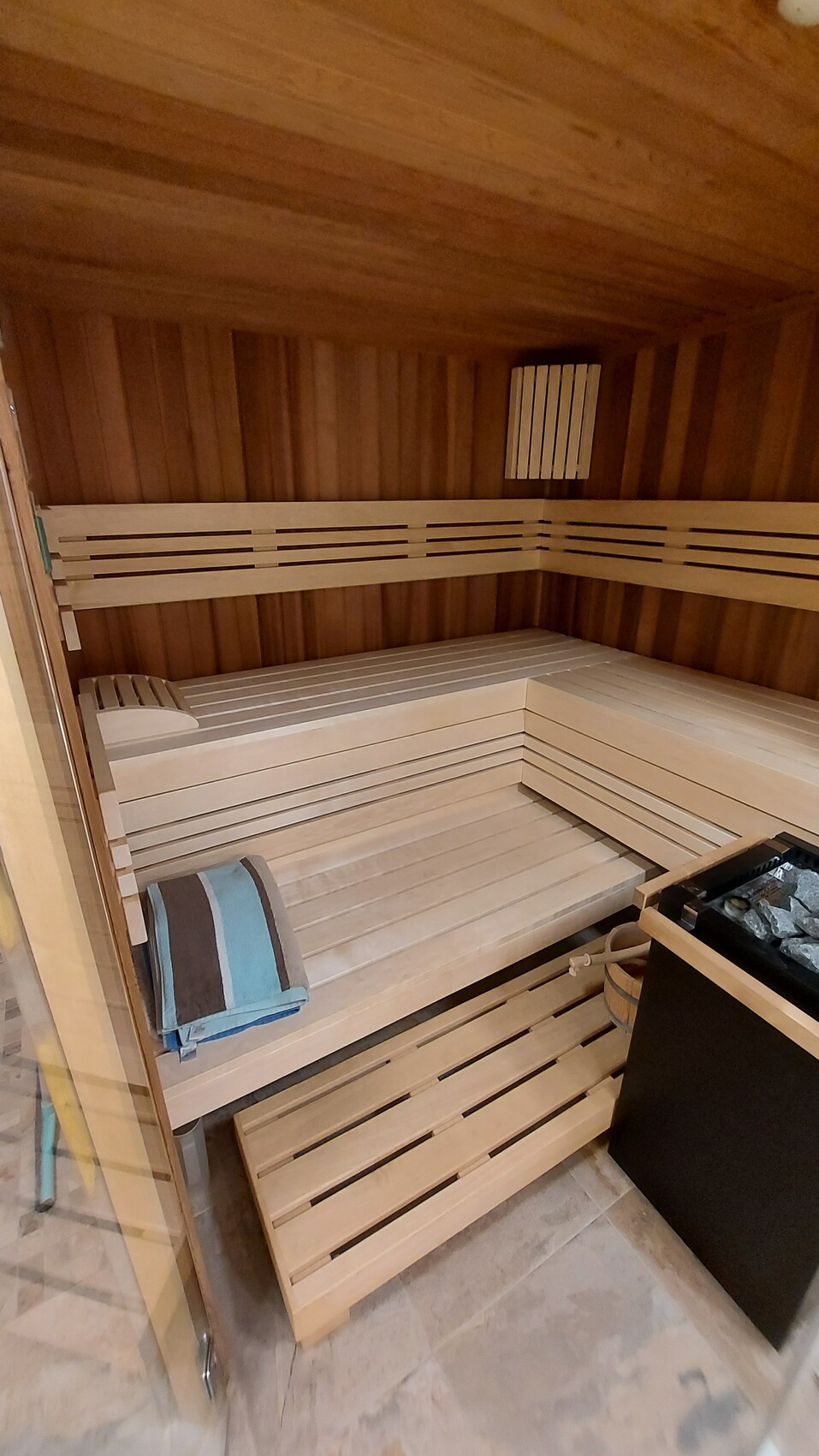 Sauna mit zwei Holzbankreihen in L-Form, Ofen mit Steinen, Holzverkleidung, Handtuch.