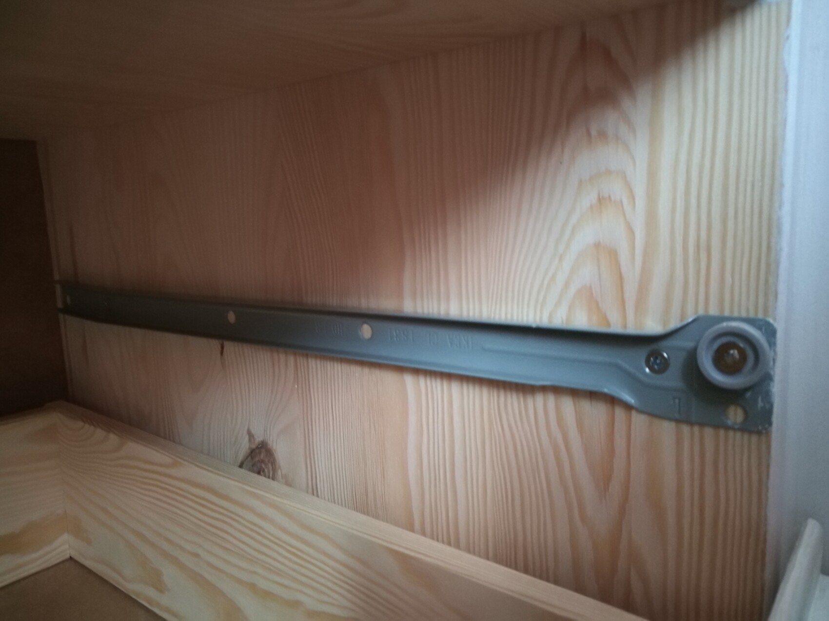 Metall-Schubladenführung an Holzwand, seitlich befestigt, mit Radrolle am rechten Ende.