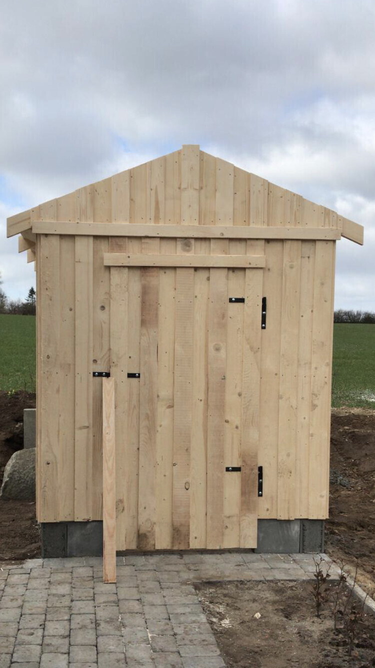 Kleiner Holzschuppen aus vertikalen Brettern mit Tür rechts, auf Betonfundament im Freien.