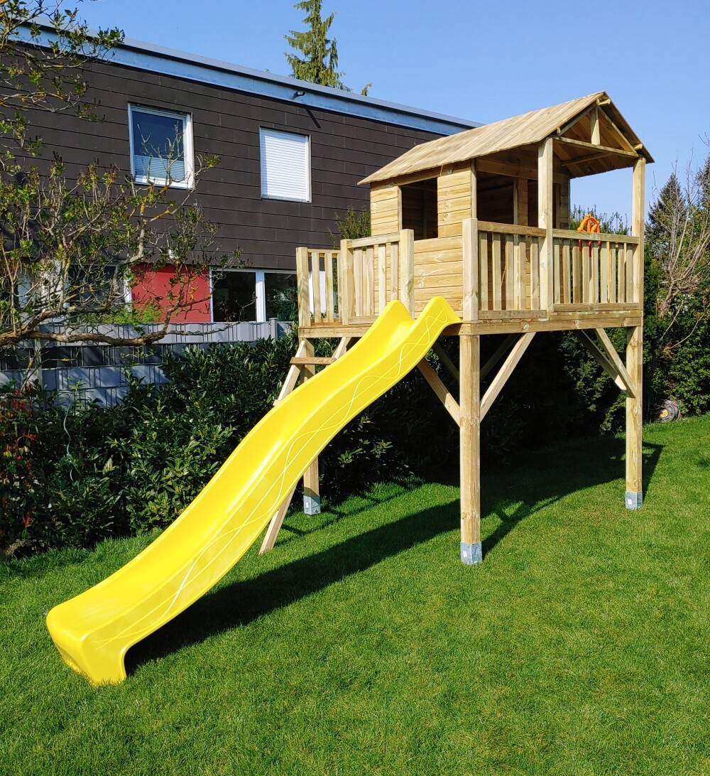 Holz-Spielturm mit gelber Rutsche auf Stelzen im Garten vor dem Haus.