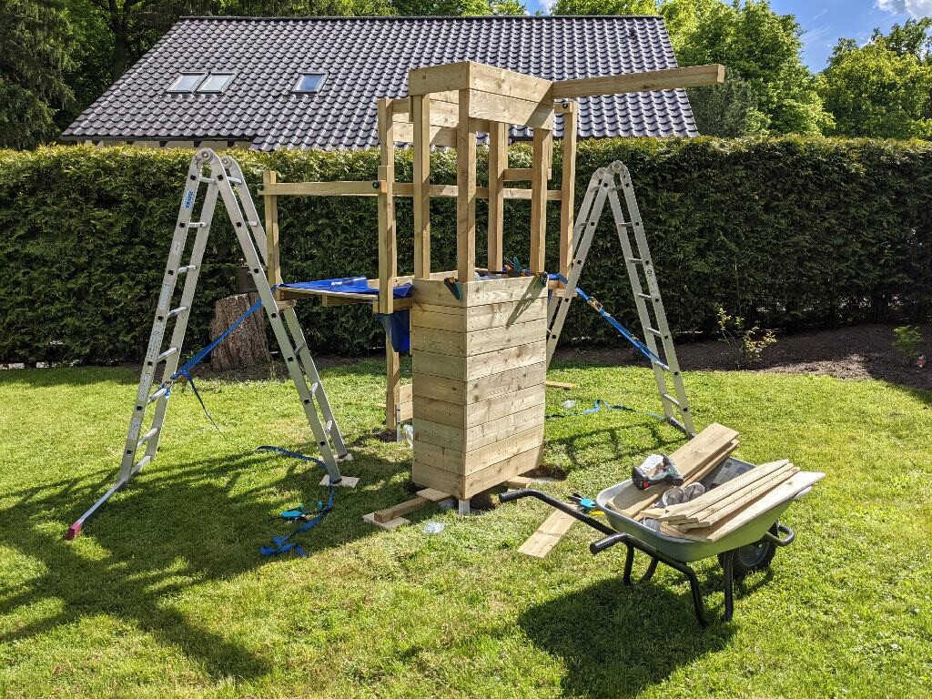 Holz-Spielgerüst im Garten im Aufbau, Leitern seitlich, Schubkarre voller Holzteile.