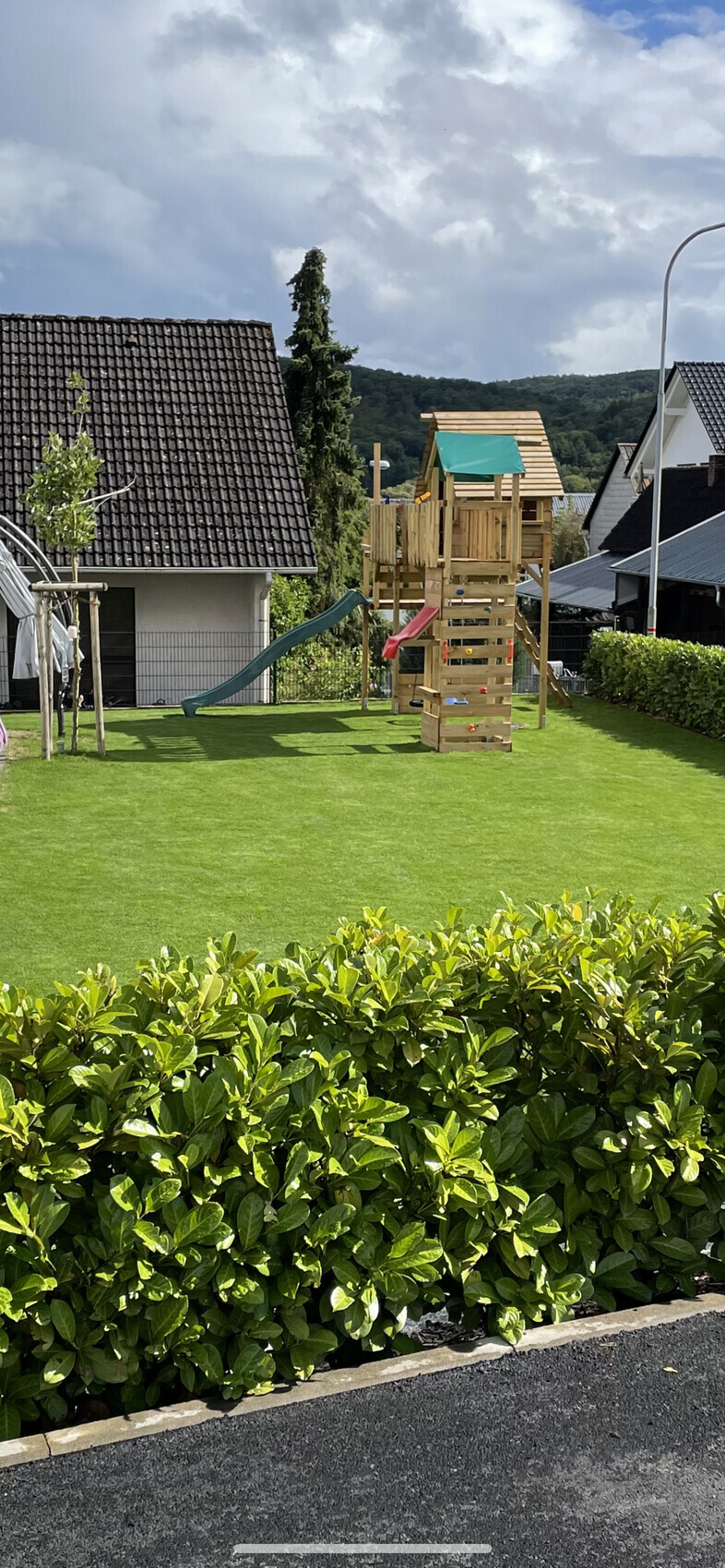 Garten mit Holz-Spielturm, grüne Rutsche, gepflegter Rasen und Hecke; Häuser im Hintergrund.