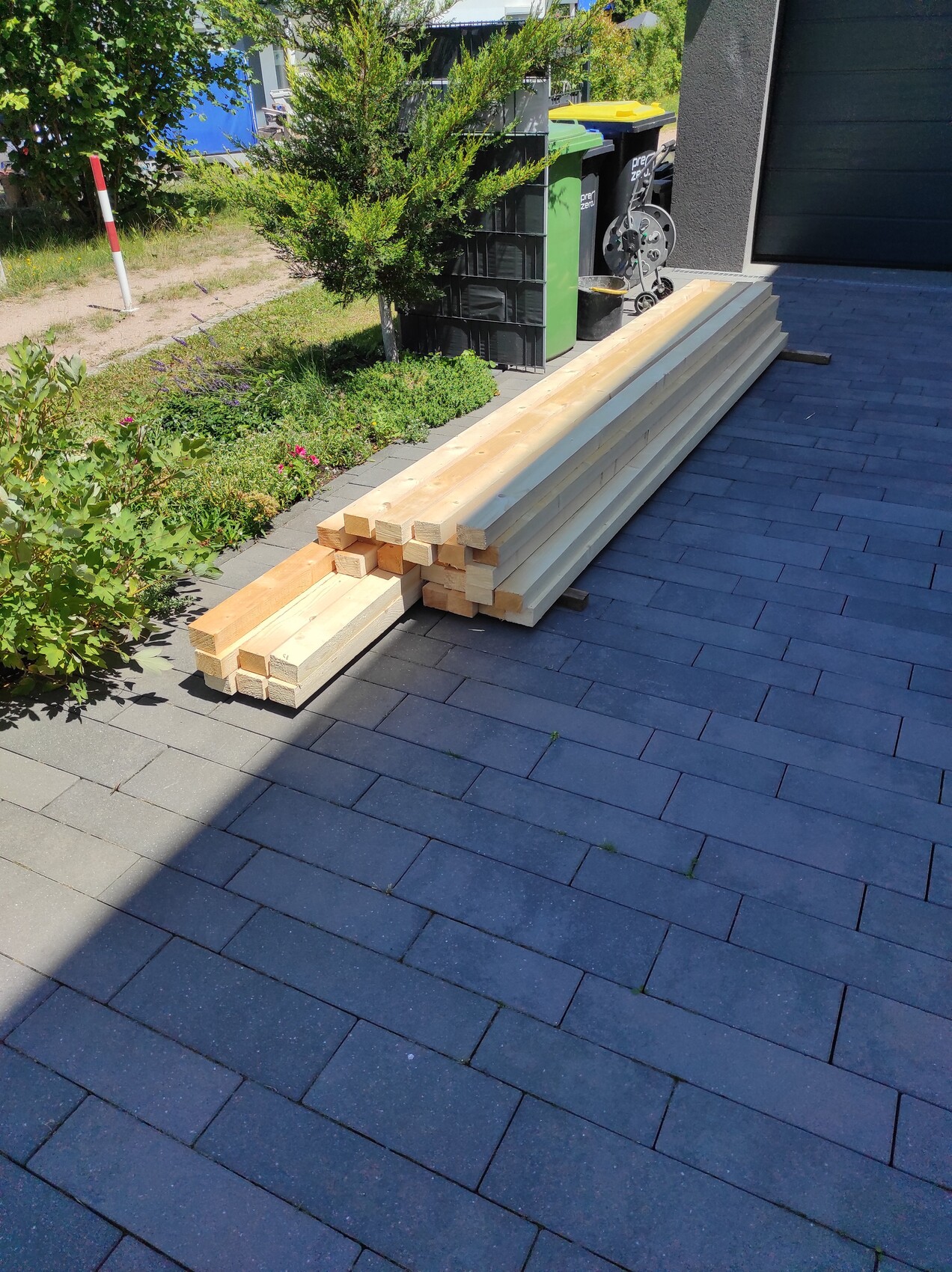 Stapel Holzlatten auf dunkler Pflasterfläche neben Garten und Mülltonnen vor einem Haus