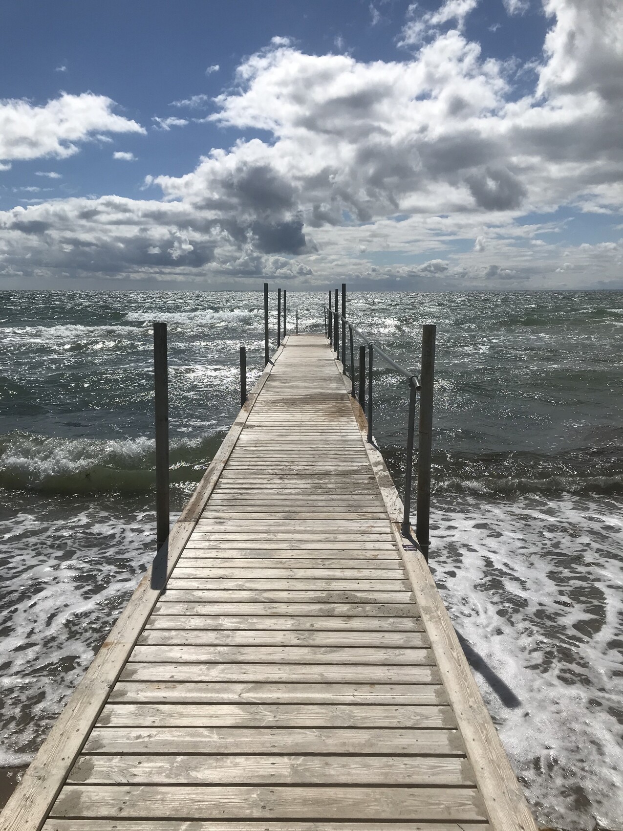 Holzsteg ins Meer mit Geländerpfosten, Wellen schlagen an den Strand, Wolkenhimmel.
