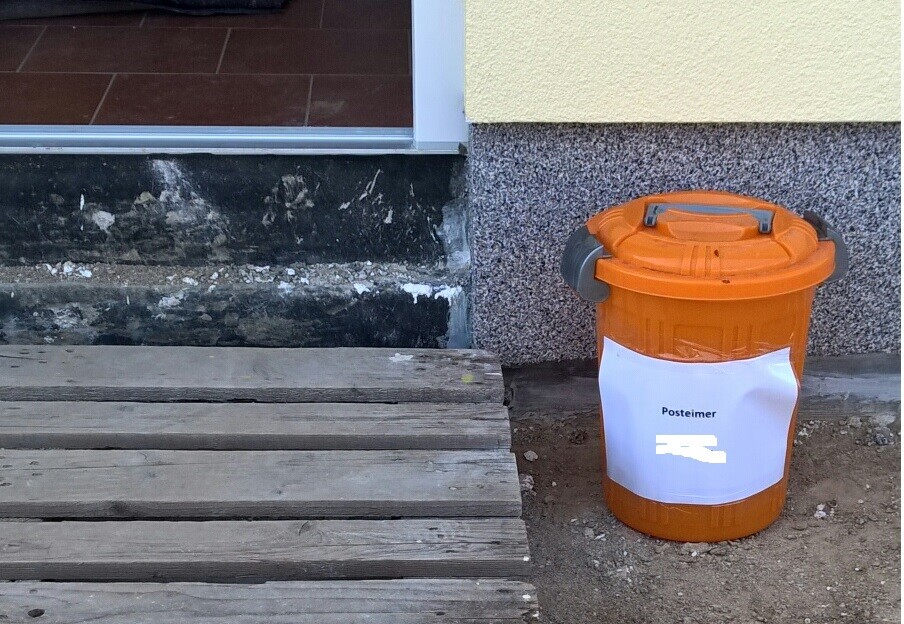 Holzstufen führen zum Eingang eines Hauses, daneben steht ein orangefarbener Müllbehälter.
