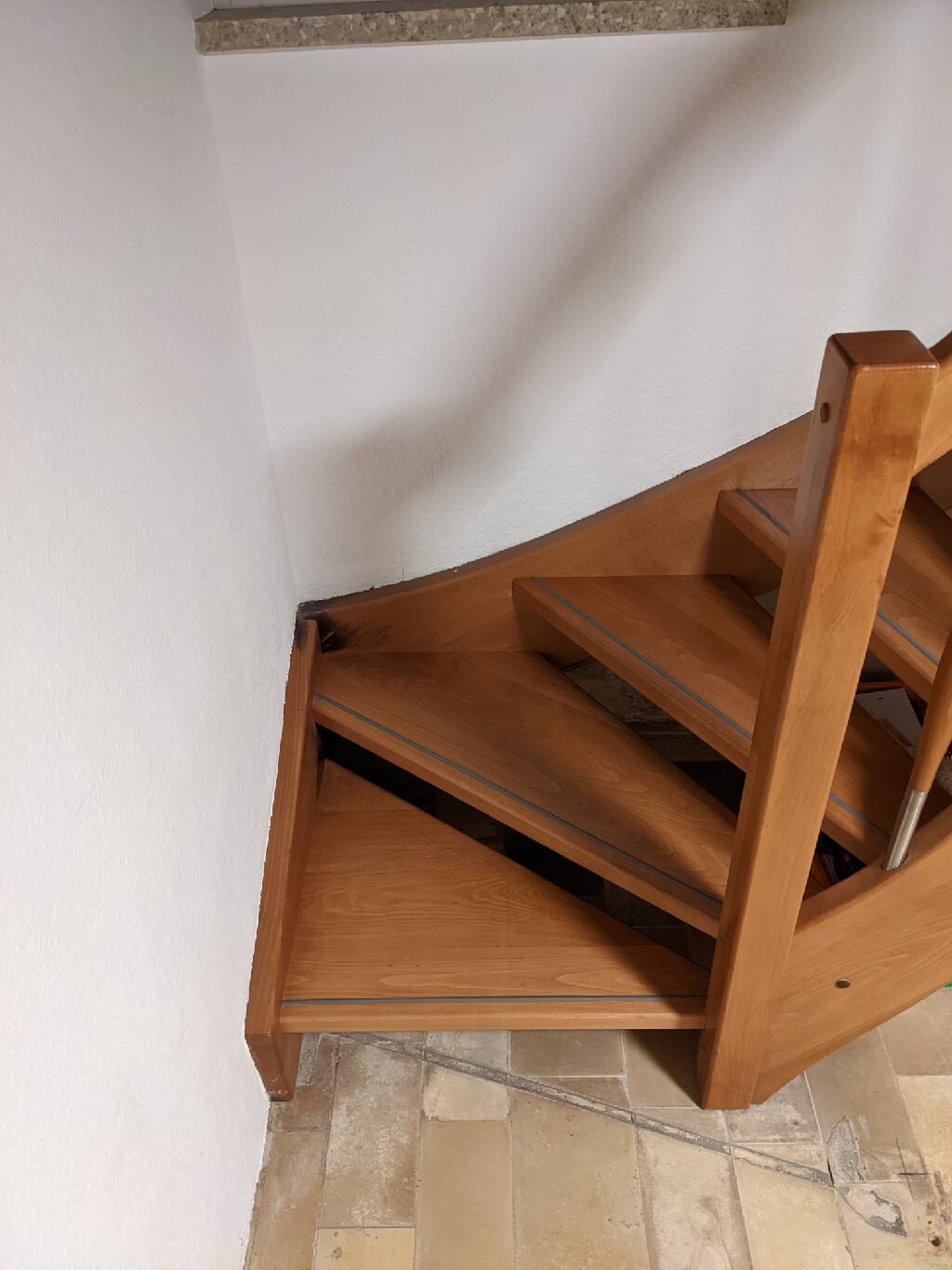 Holztreppe mit Geländer im Innenraum, Eckbereich an weißer Wand, Fliesenboden sichtbar.