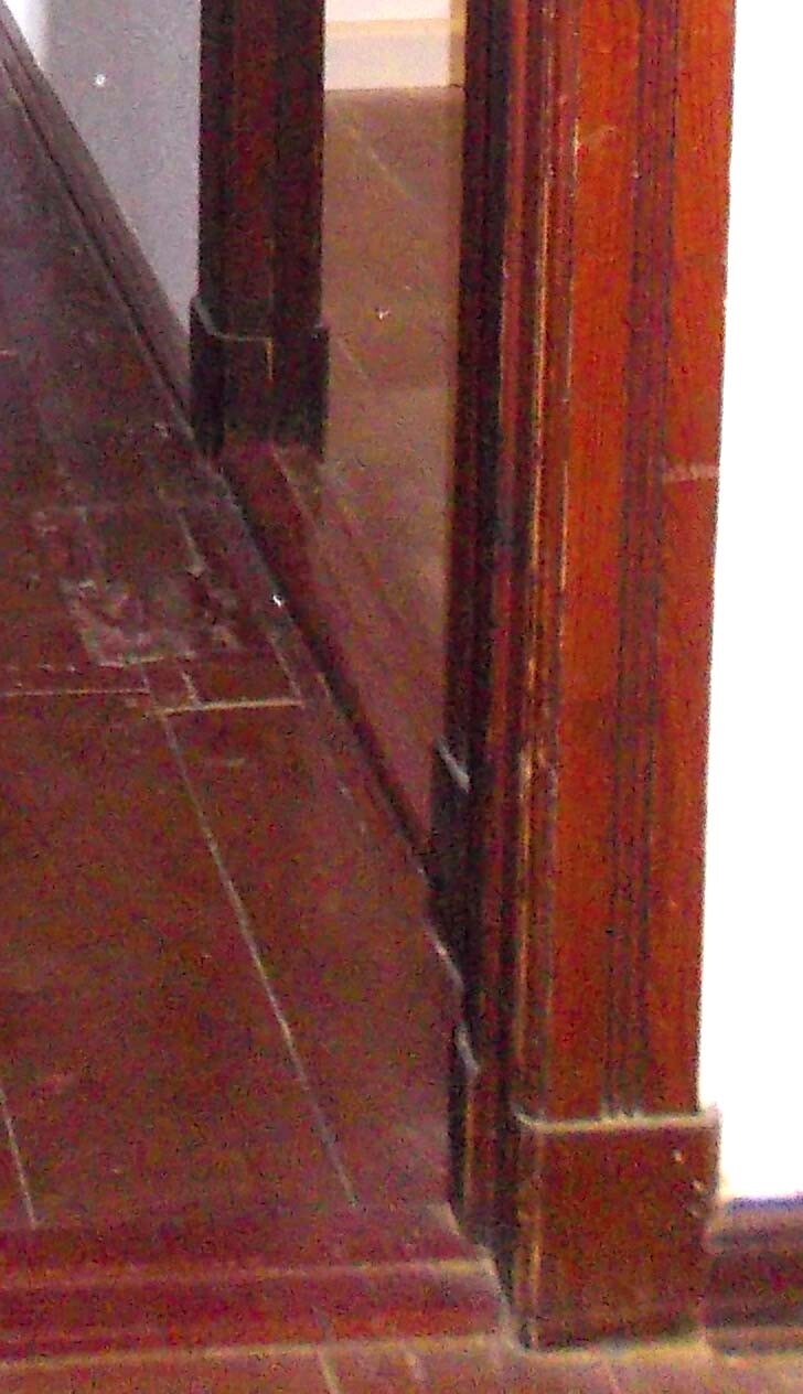 Innenraum-Ecke mit dunkler Holzwand, rechter Holz-Türrahmen, brauner Boden.