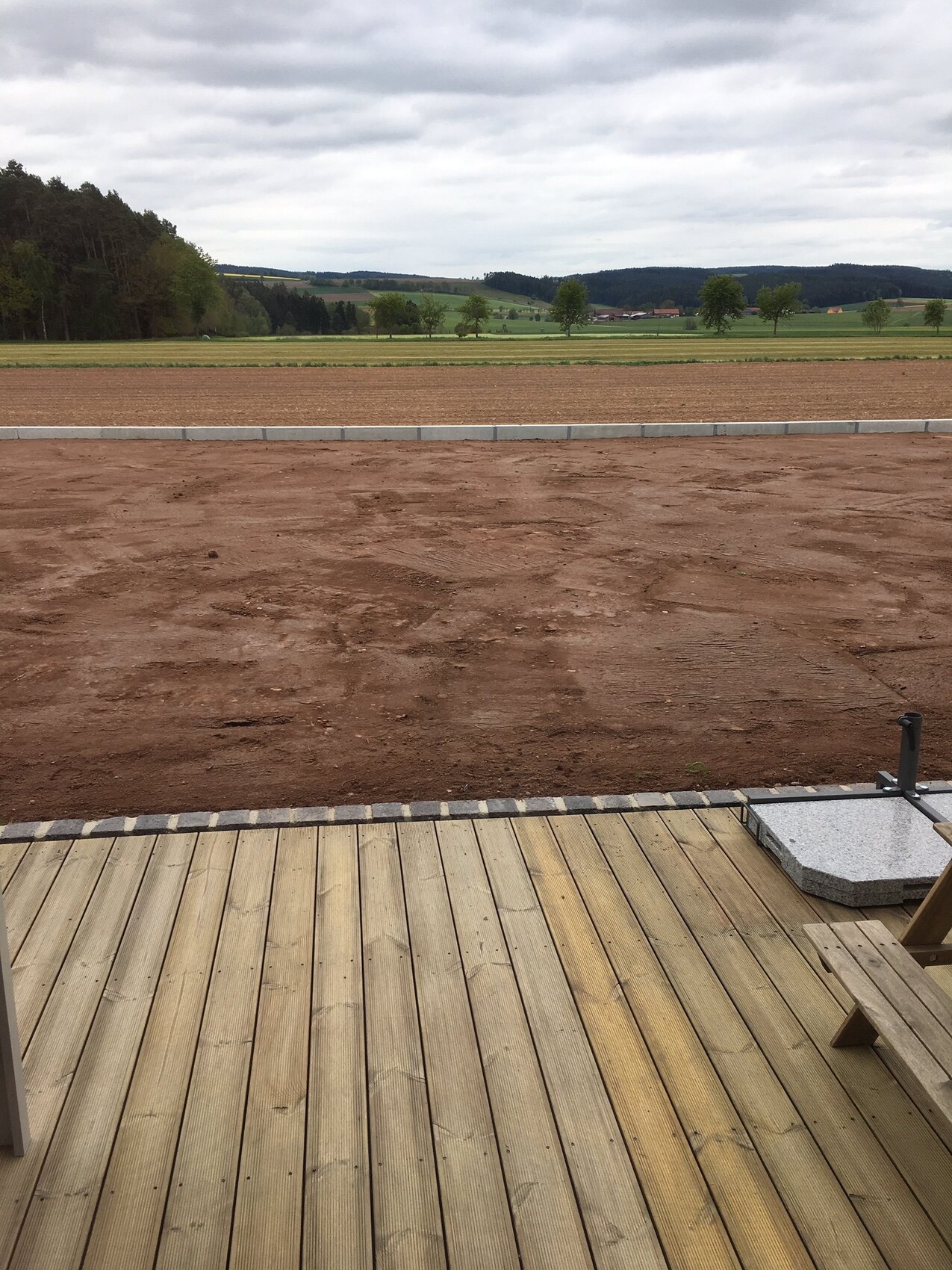Blick von einer Holzterrasse auf eine braune Erdfläche vor Felder, Bäumen und Hügeln.