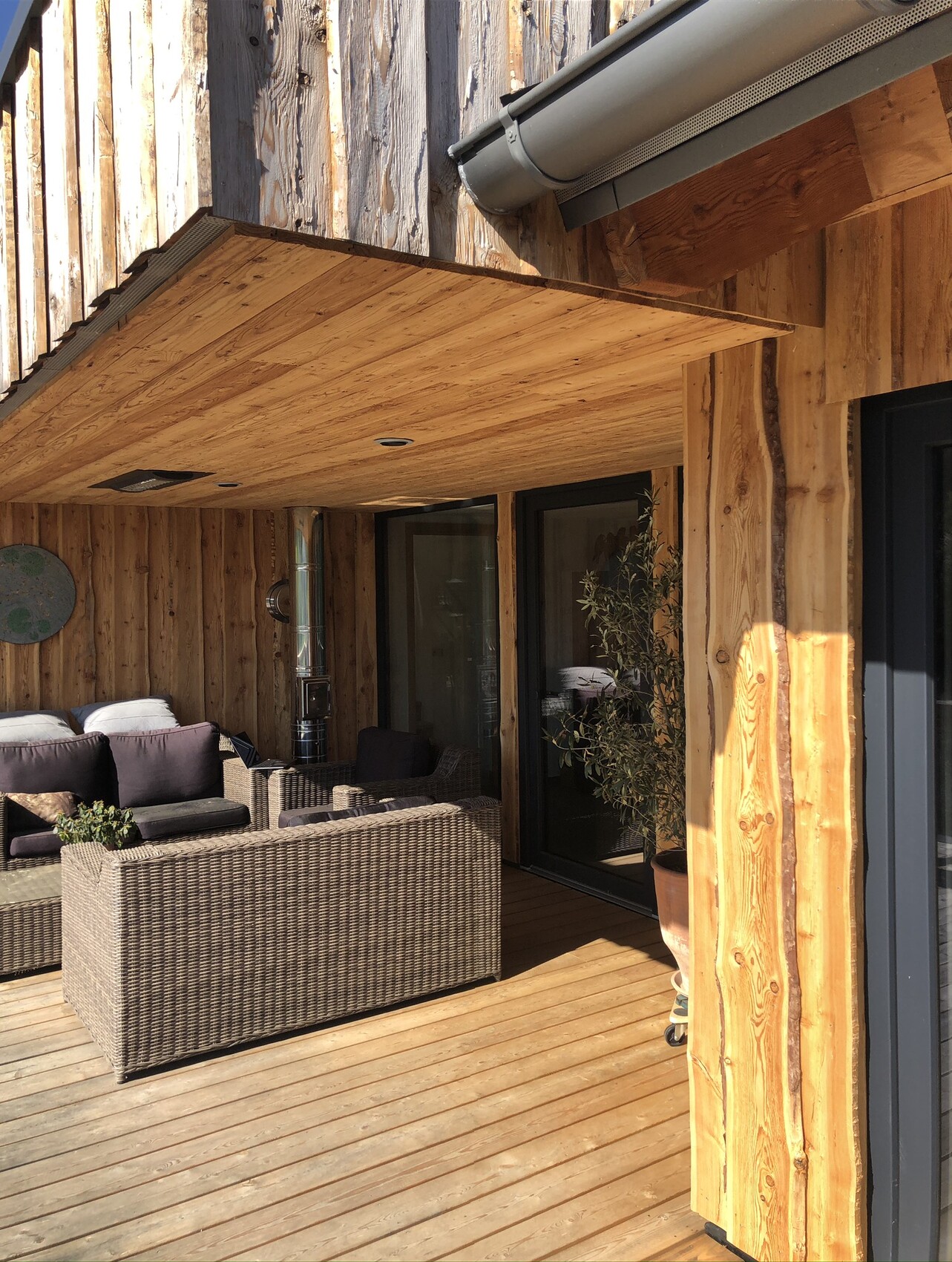 Holzterrasse mit Korbsofa-Set, Kissen, Glas-Schiebetüren und Pflanzkübel.