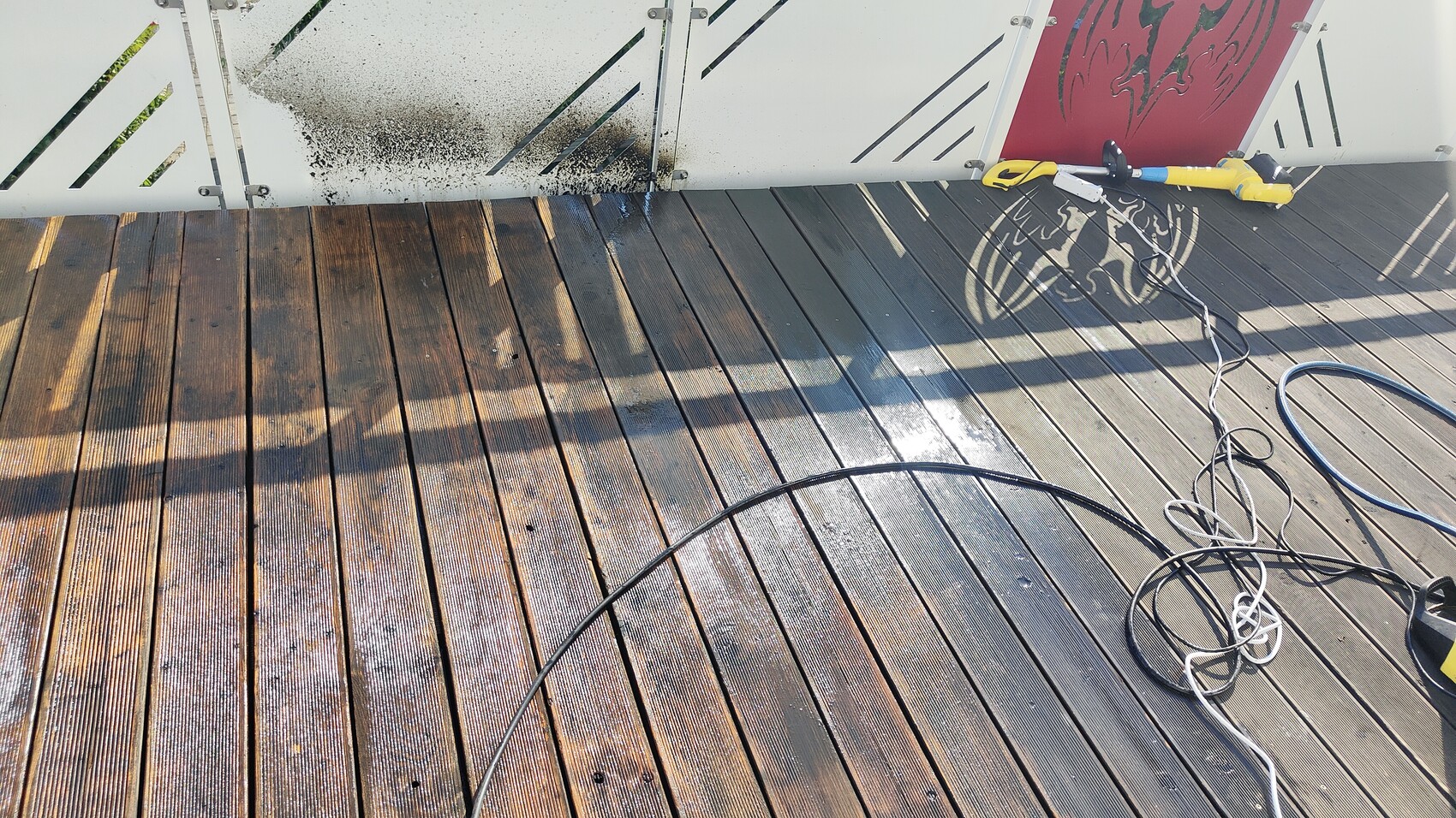 Nass Holzdielen-Terrasse; Schlauchkabel, Hochdruckreiniger am Rand, rote grafische Wand.