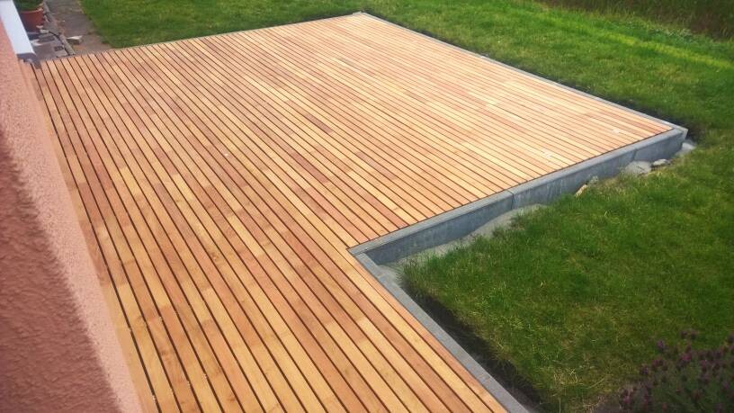 Holzterrasse aus parallel verlaufenden Planken neben grünem Rasen und Betonrand.