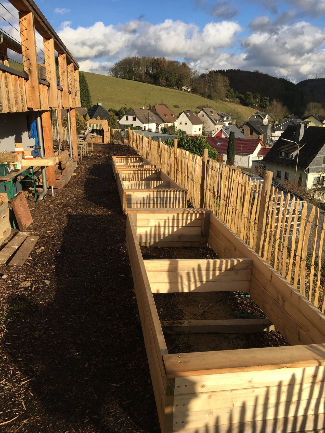 Holzterrasse mit Hochbeeten aus Holz, Blick auf dörfliche Häuser und grüne Hügel im Hintergrund.