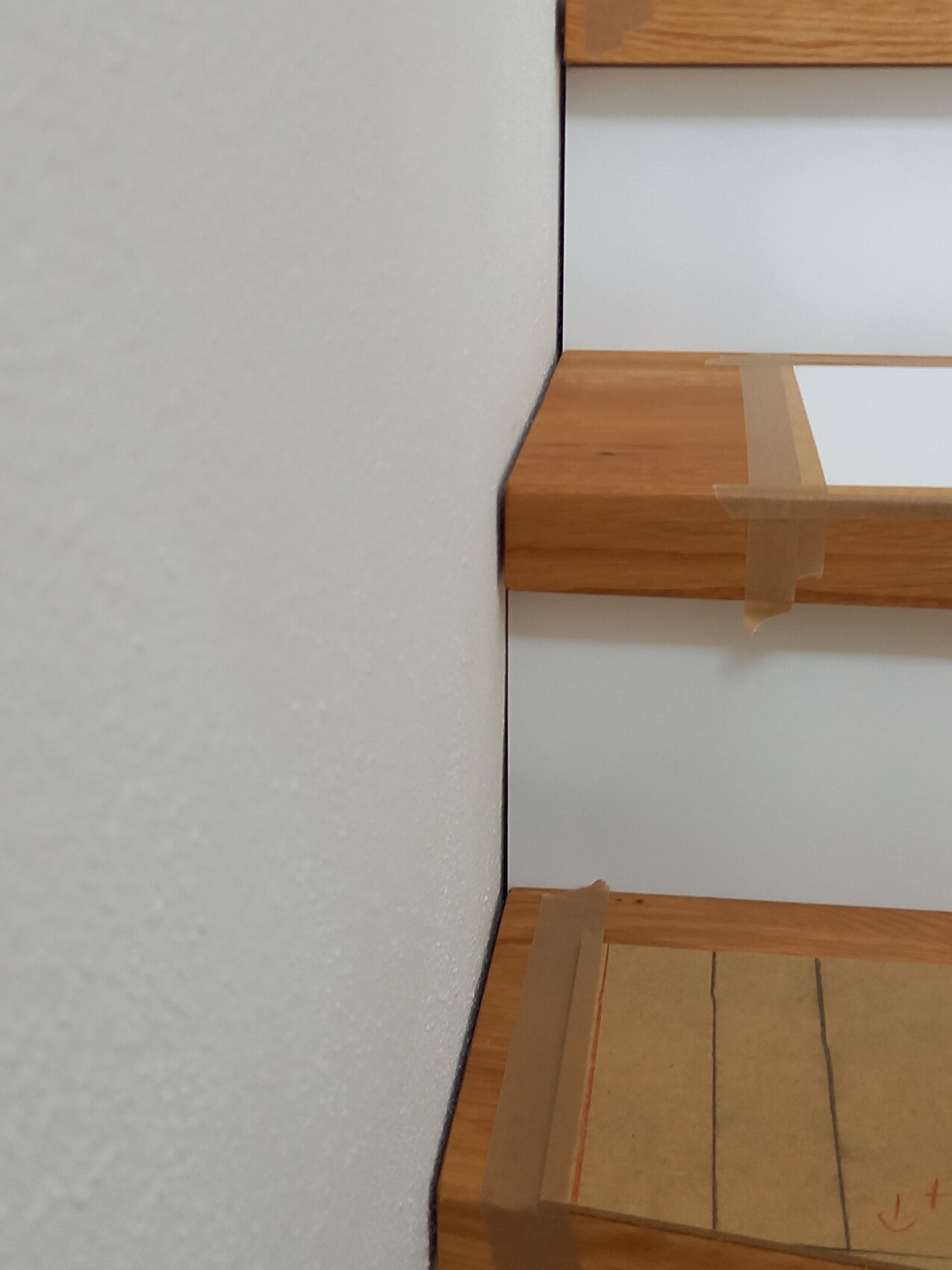 Holztreppe mit Klebeband an den Kanten; Kartonabdeckung auf der unteren Stufe; graue Wand links.