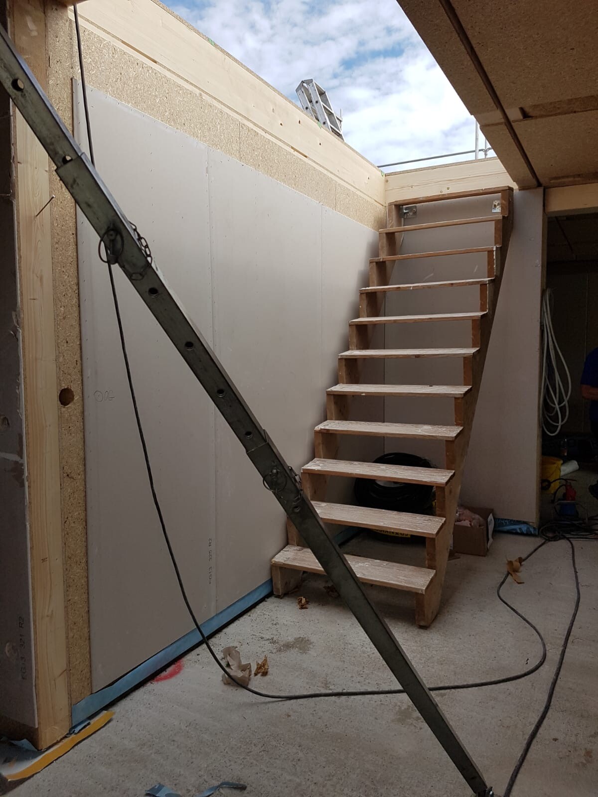 Holztreppe im Rohbau neben einer diagonal verlaufenden Metallstrebe; Kellergeschoß mit Kabeln