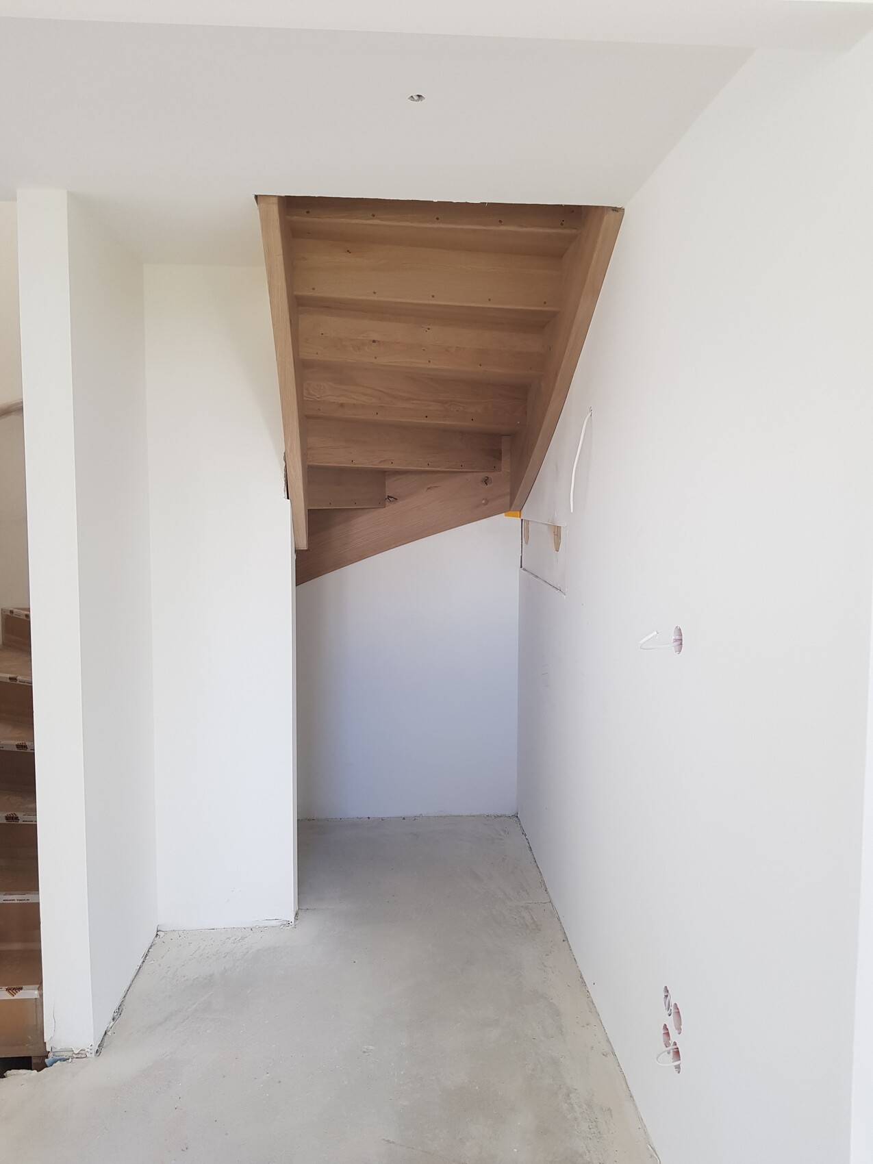 Holztreppe mit sichtbaren Stufen führt nach oben, darunter ein kleiner Lagerraum; weiße Wände.