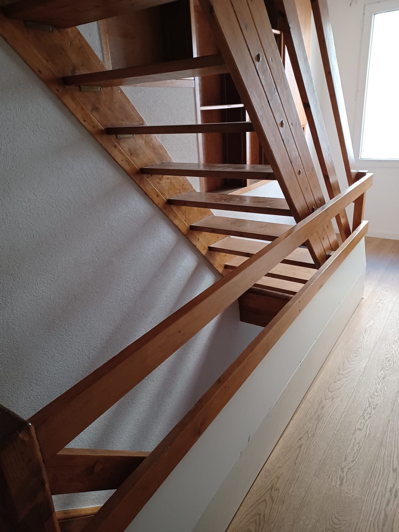 Holztreppe mit Geländer im Innenraum, vor heller Wand, Fenster oben rechts.