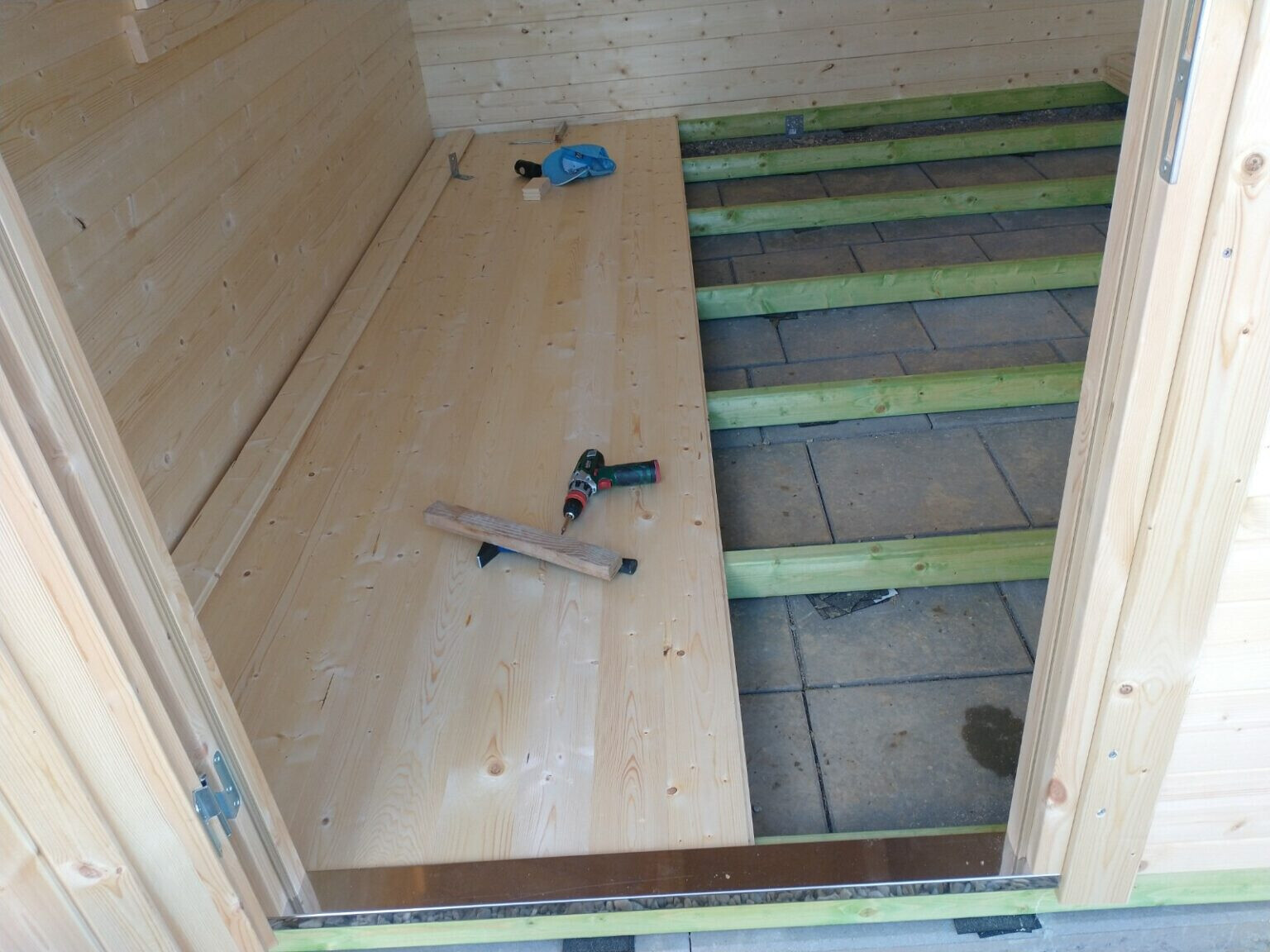 Innenraum eines Holzbaus: Bohrmaschine liegt auf dem Holzboden, grün gestrichene Stufenrahmen.