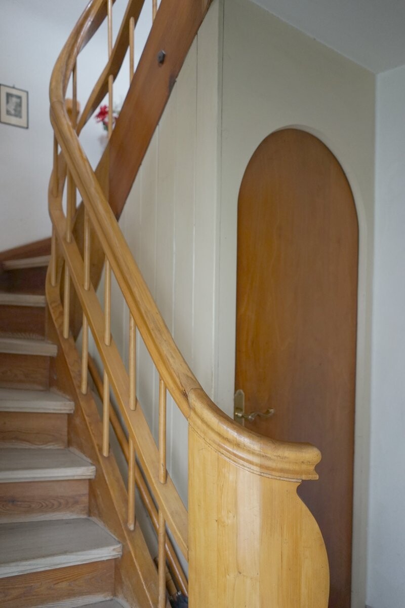Holztreppe mit Geländer, daneben eine Tür mit rundem Oberteil an der Wand.