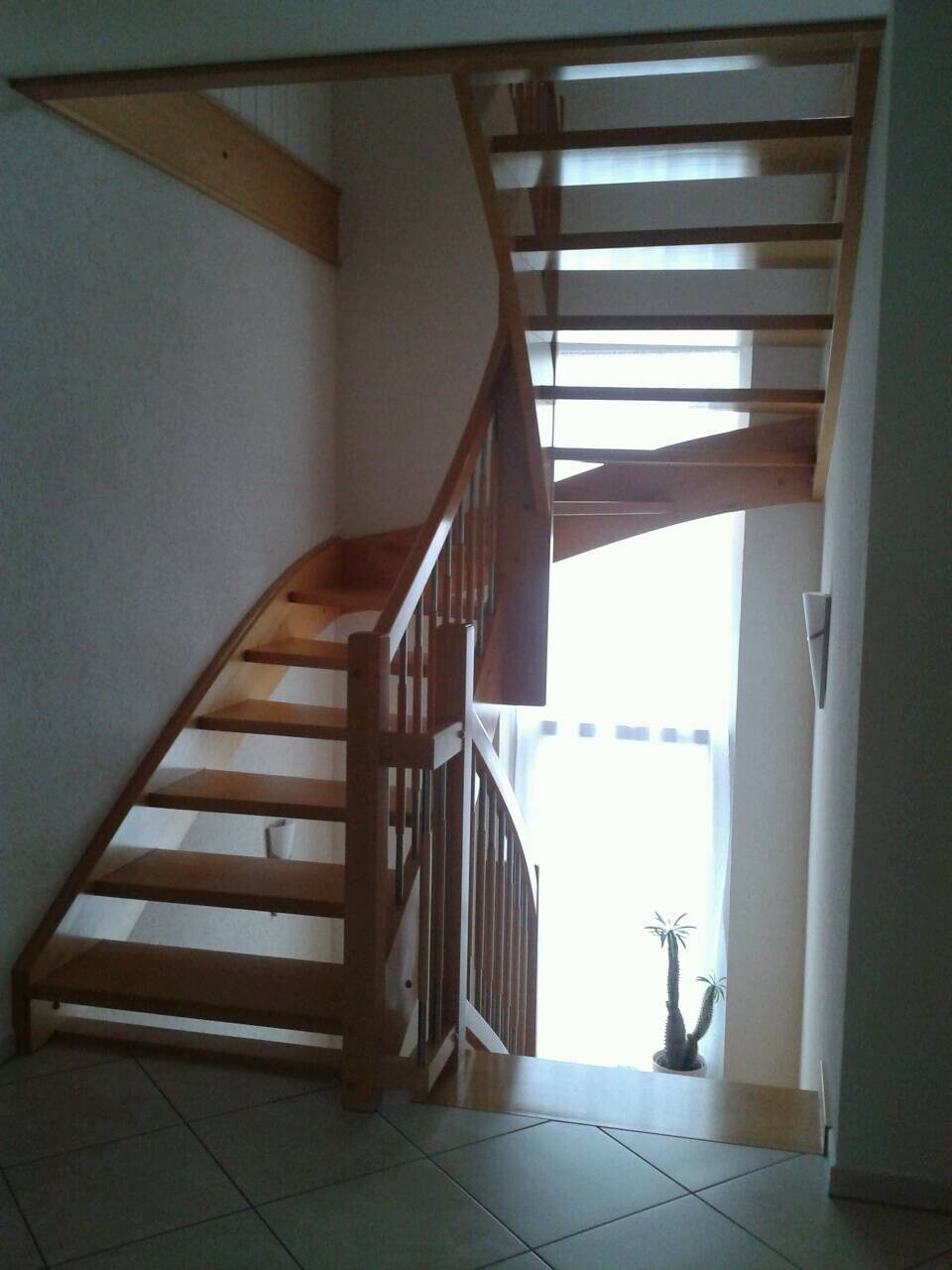 Holztreppe mit offenen Stufen und Geländer, Fenster dahinter, Pflanze rechts.