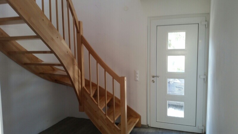Innenraum mit gebogener Holztreppe links und weißer Tür rechts mit Glasfenstern.