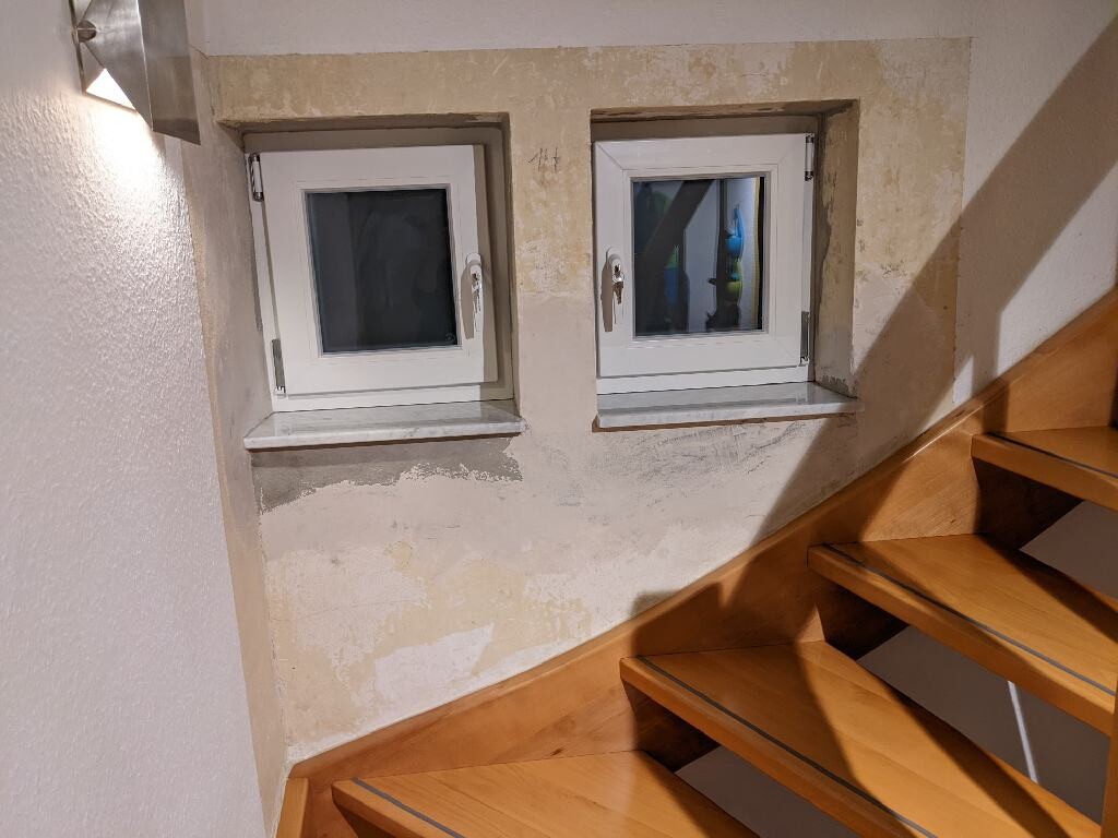 Zwei kleine Fenster in einer beschädigten, verputzten Wand neben einer Holztreppe im Innenraum.