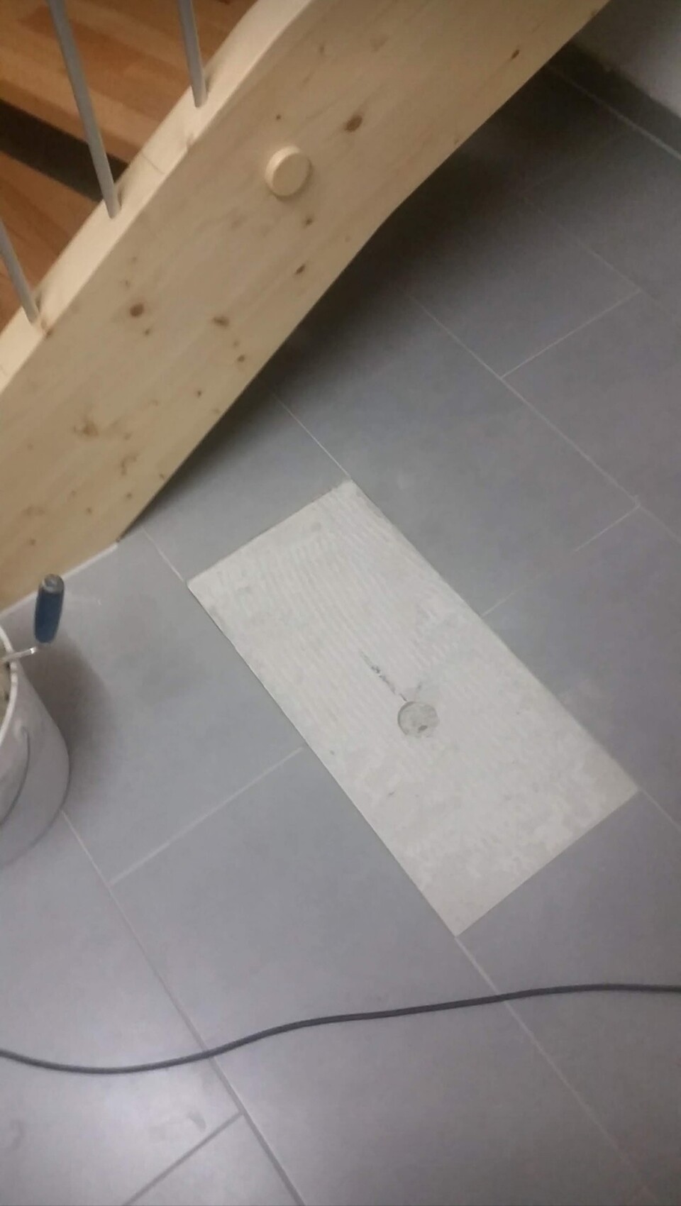 Grauer Fliesenboden mit rechteckiger Reparaturfläche und rundem Deckel; Eimer und Kabel daneben.