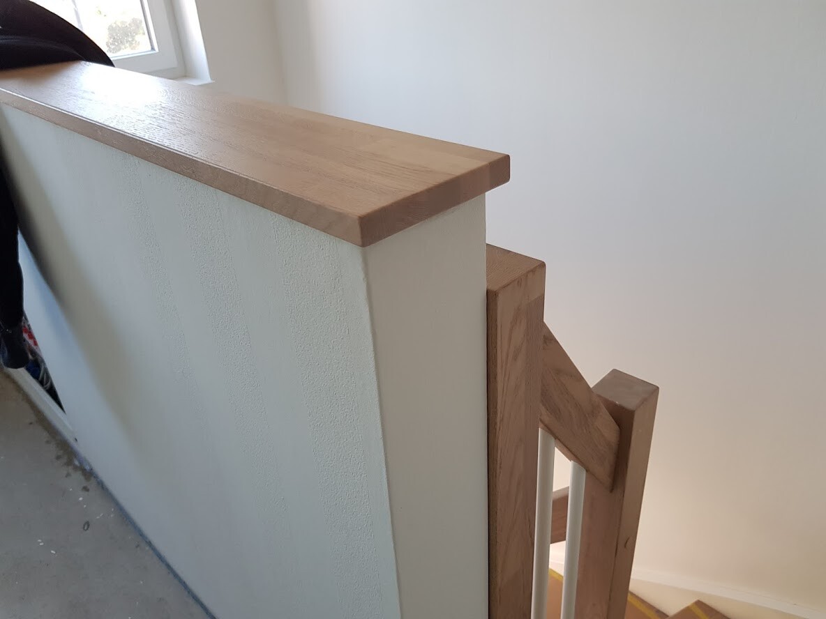 Holzhandlauf und Geländer an weißer Treppenbrüstung neben Treppenstufen, Fenster links.