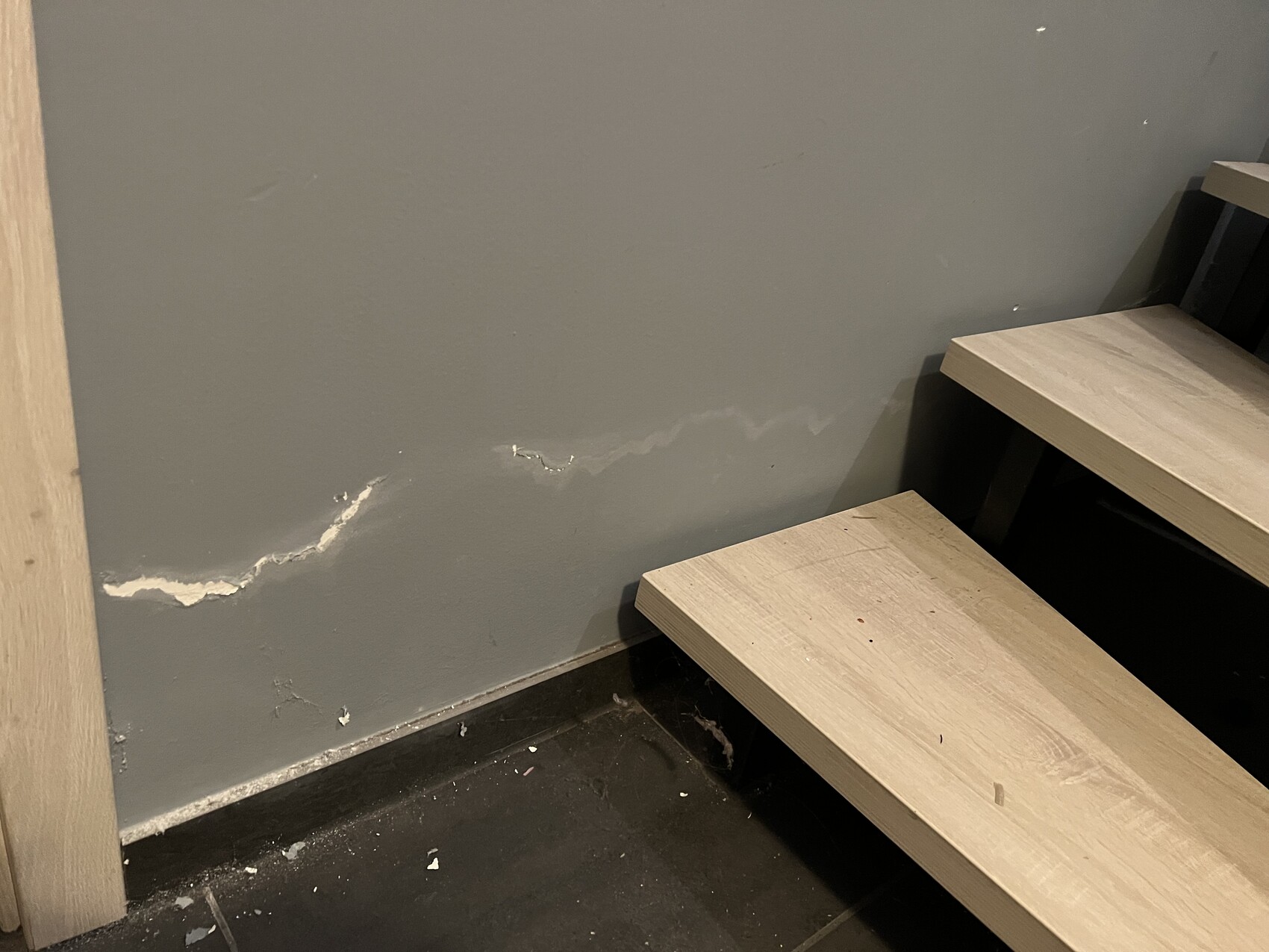 Graue Wand mit Rissen und Putzabplatzungen am unteren Rand neben hellen Holzstufen