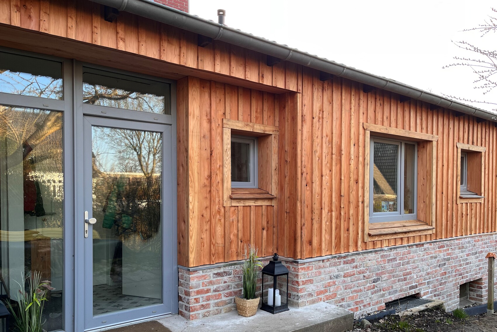 Holzverkleidete Außenwand mit Glastür links und mehreren Fenstern, Steinfundament.