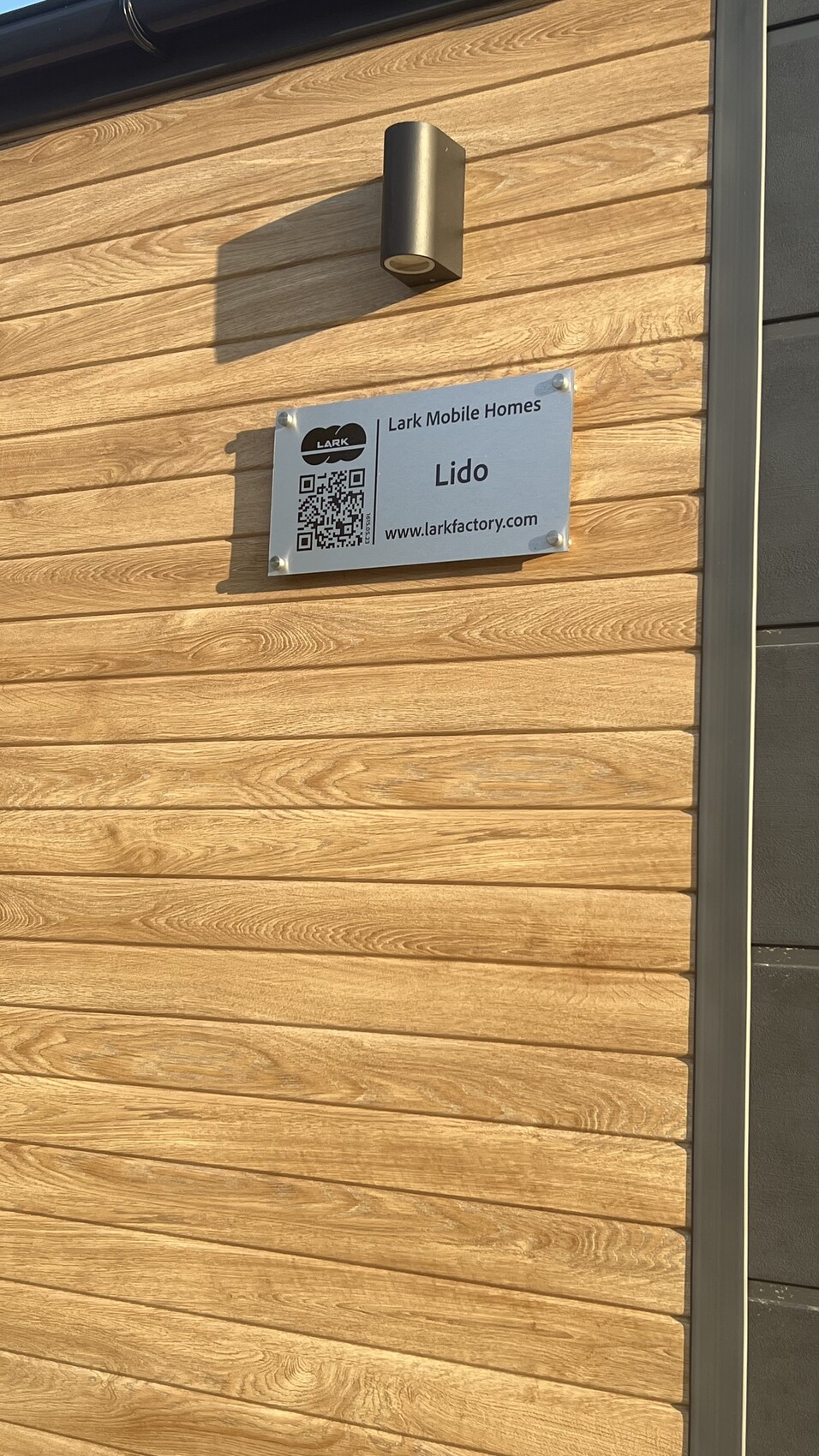 Holzverkleidung einer Außenwand mit Metall-Schild Lark Mobile Homes Lido, QR-Code und Außenleuchte