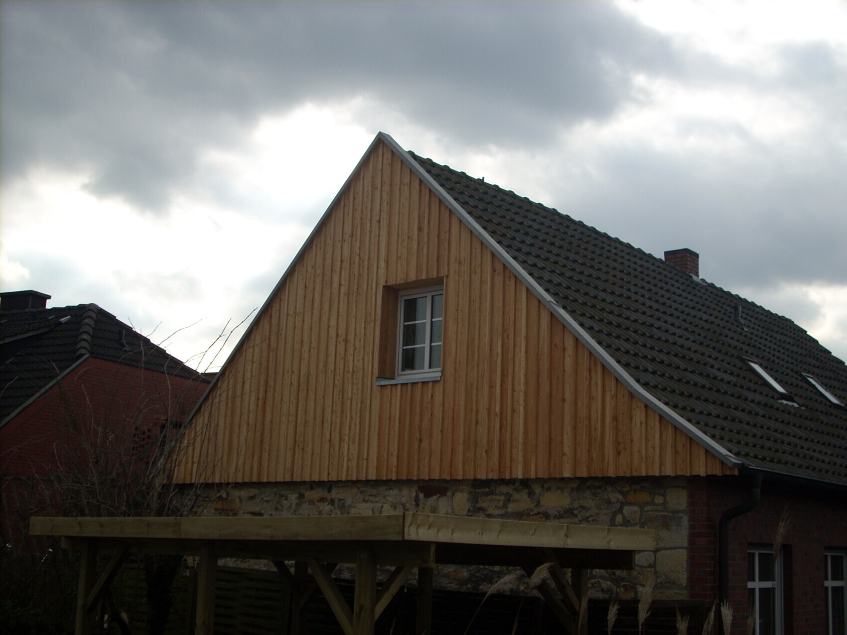 Holzverkleidetes Haus mit schrägem Giebel, Steinunterbau und grauem Dach vor Wolkenhimmel.