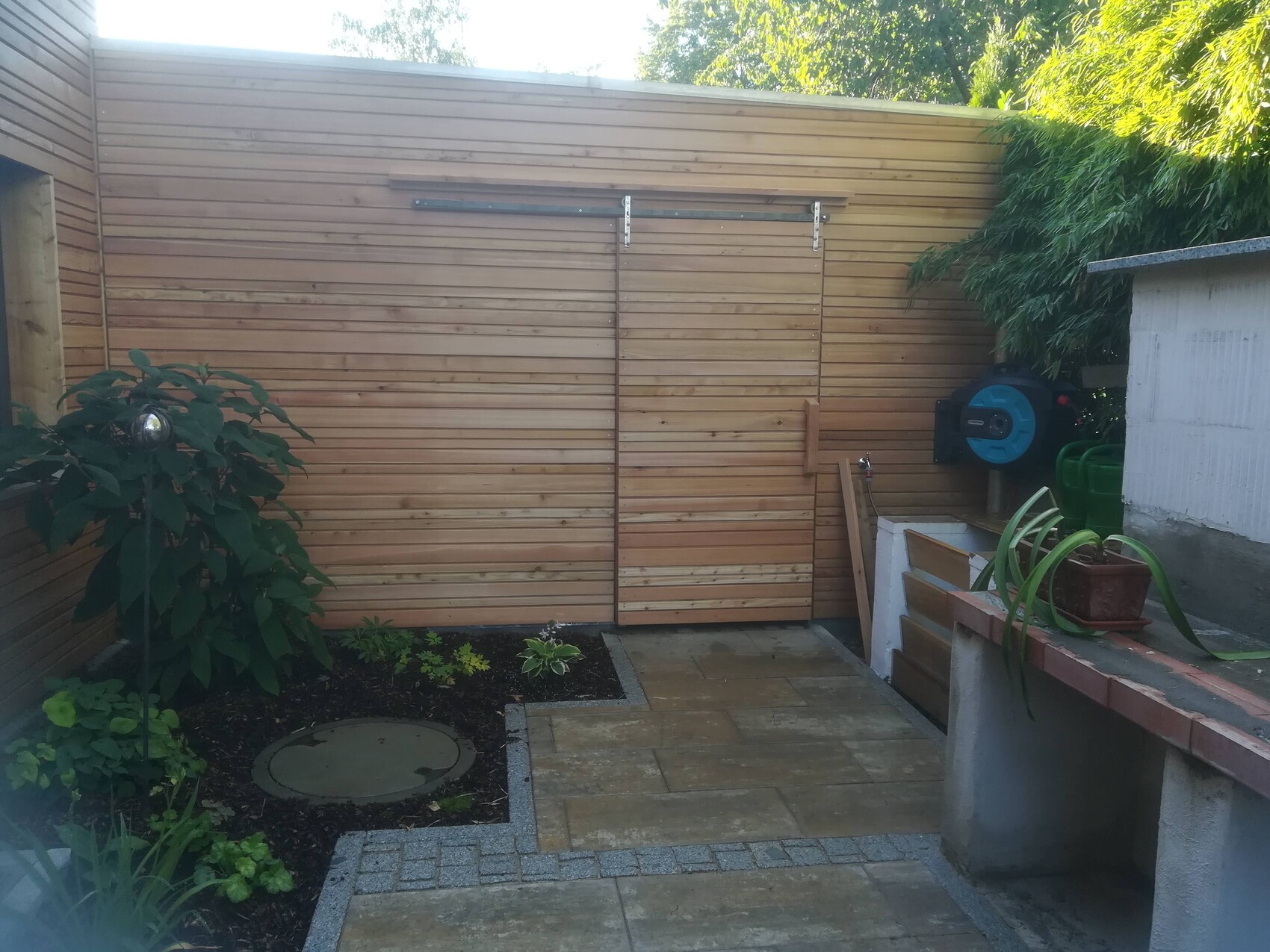 Gartenbereich mit Holzlattenwand, Pflanze links, Wasserhahn, Schlauchaufroller rechts.